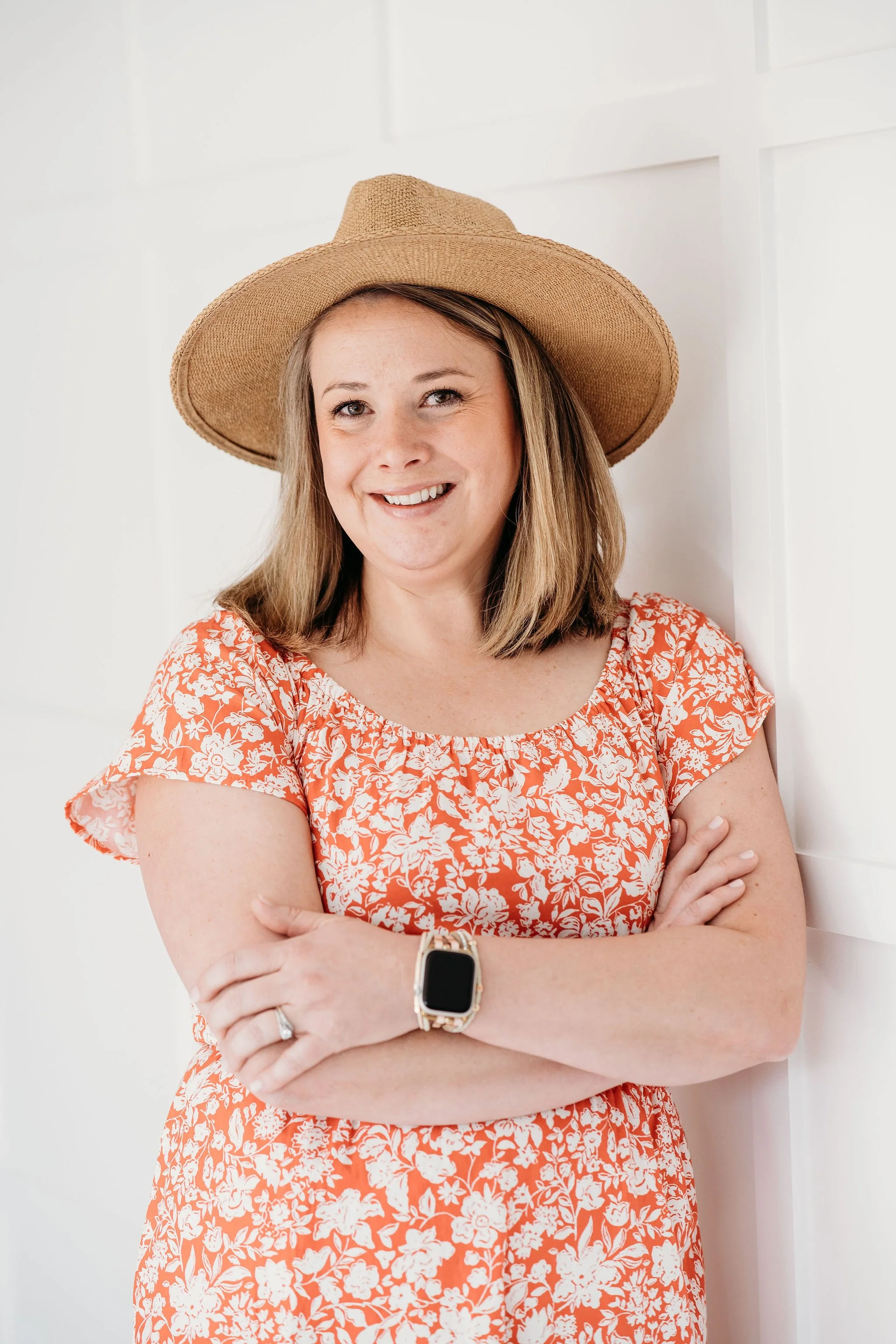 Woman wearing a straw hat, orange floral dress, and smartwatch, standing with arms crossed and smiling against a white-paneled wall.