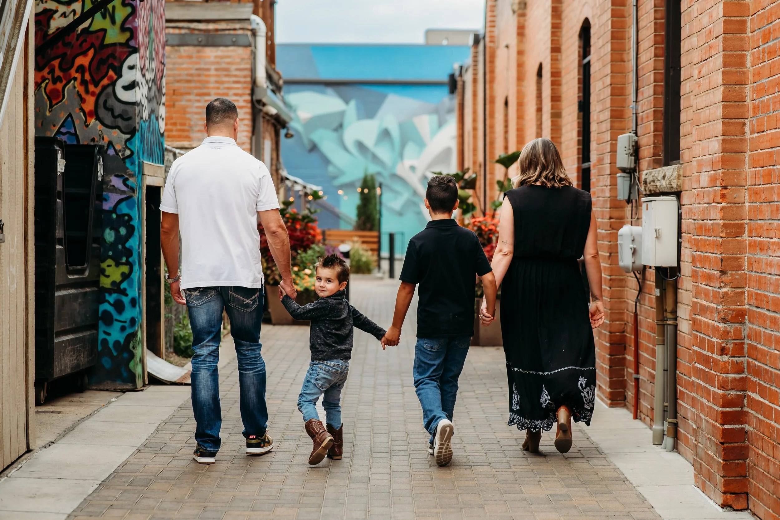 A family of four walking down a brick alleyway, holding hands. The family includes a man, a woman, and two young boys, one of whom is looking back at the camera.