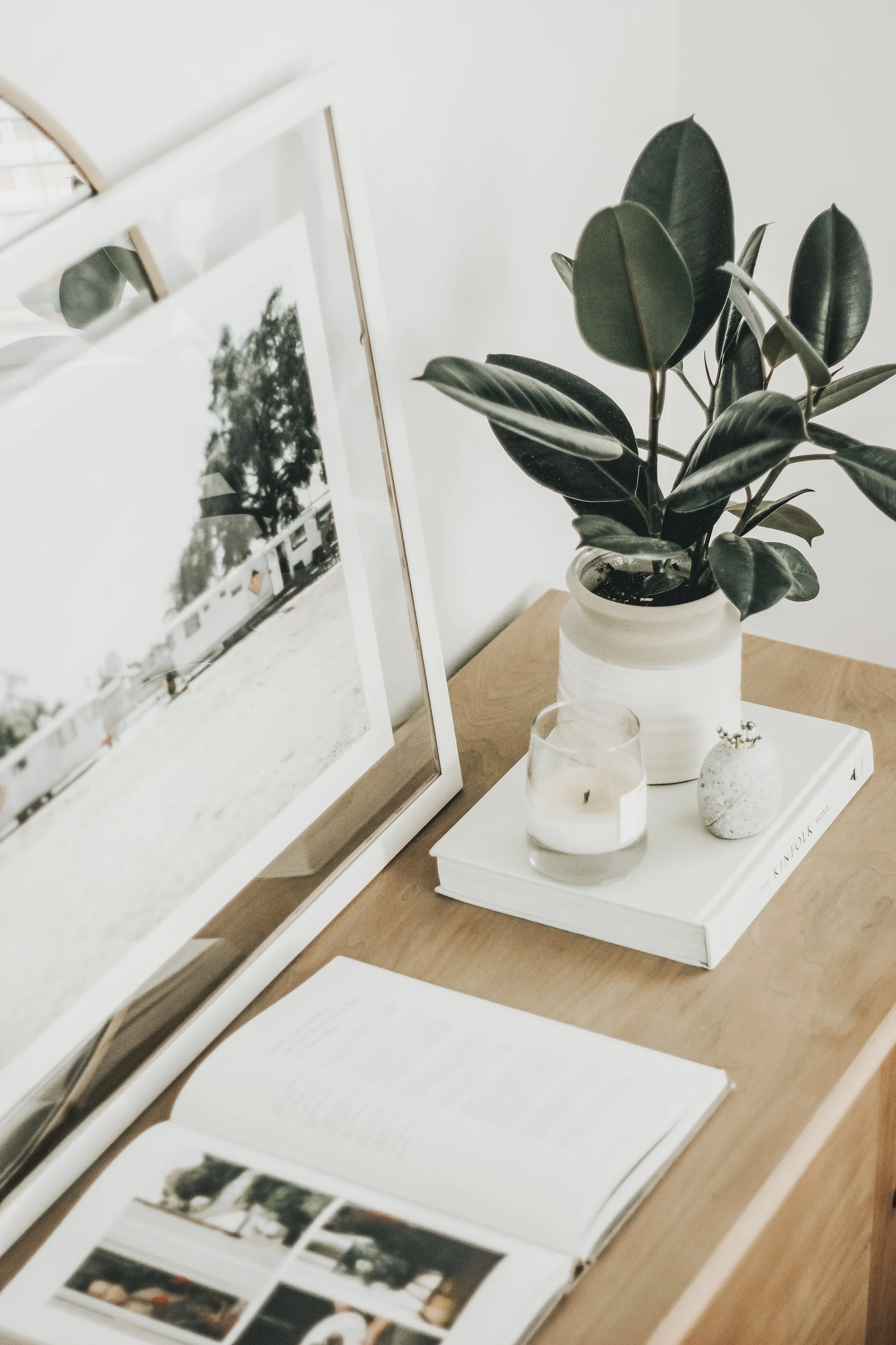 Decorative workspace with a white framed photo, potted plant, lit candle in a glass holder, small decorative stone, and a hardcover book on a wooden surface.