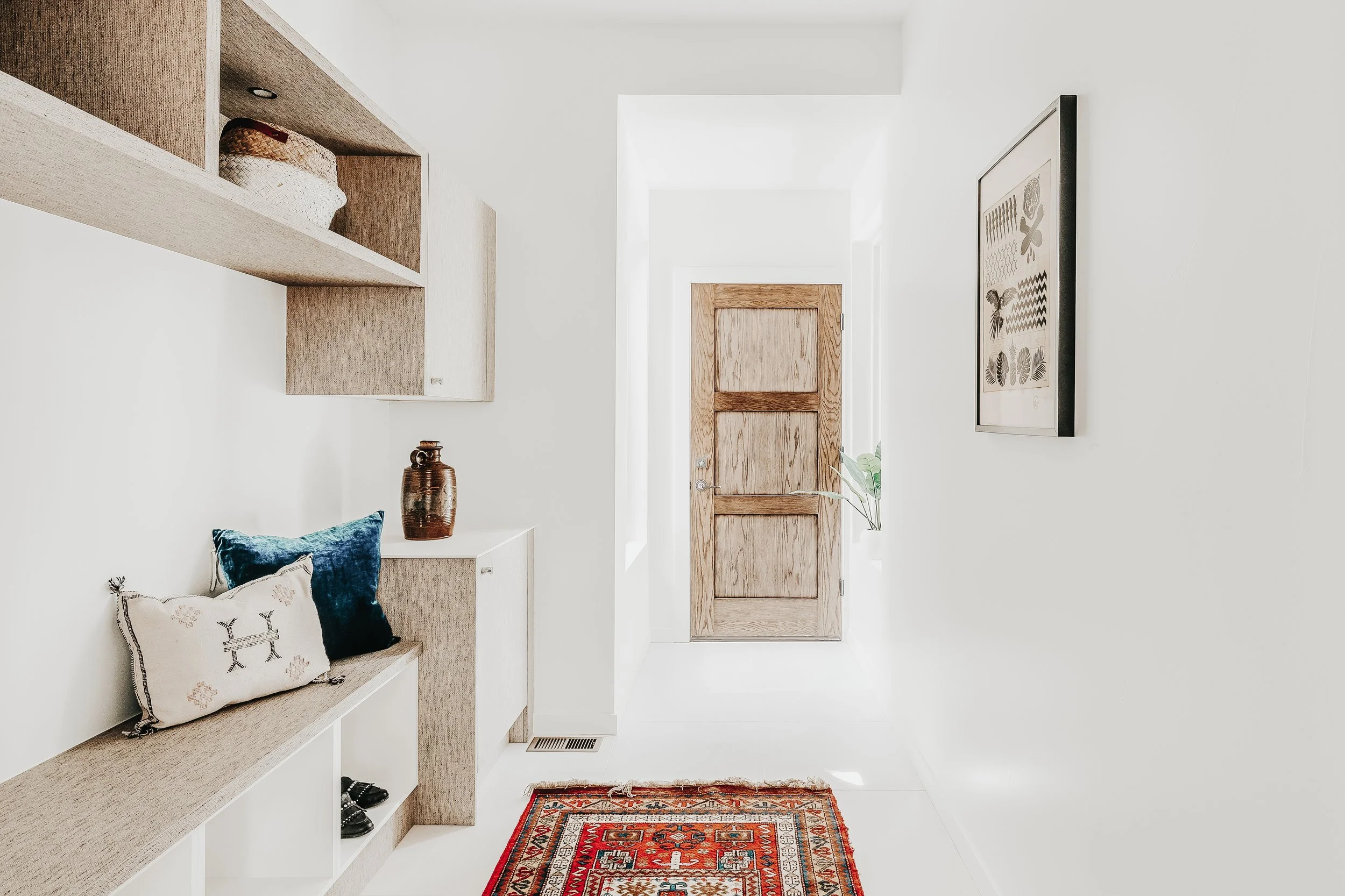 Bright, minimalist entryway with white walls, wooden door, and a decorative red oriental rug. Decor includes pillows on a built-in shelf, a brown pottery vase, and framed artwork with botanical prints.