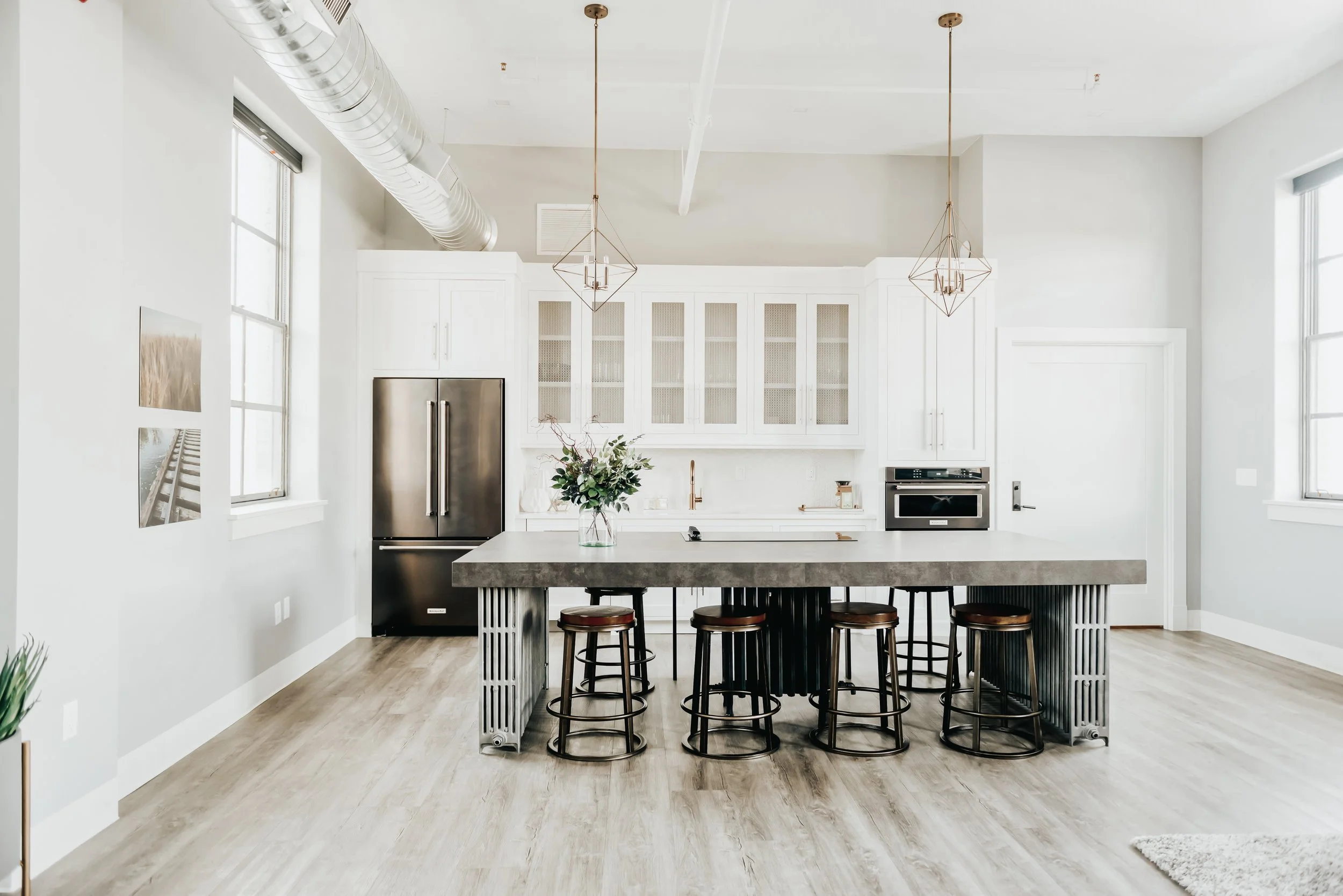 Modern kitchen with white cabinets, stainless steel refrigerator, large island with four black stools, light wood flooring, pendant lighting, and decorated with a vase of flowers.