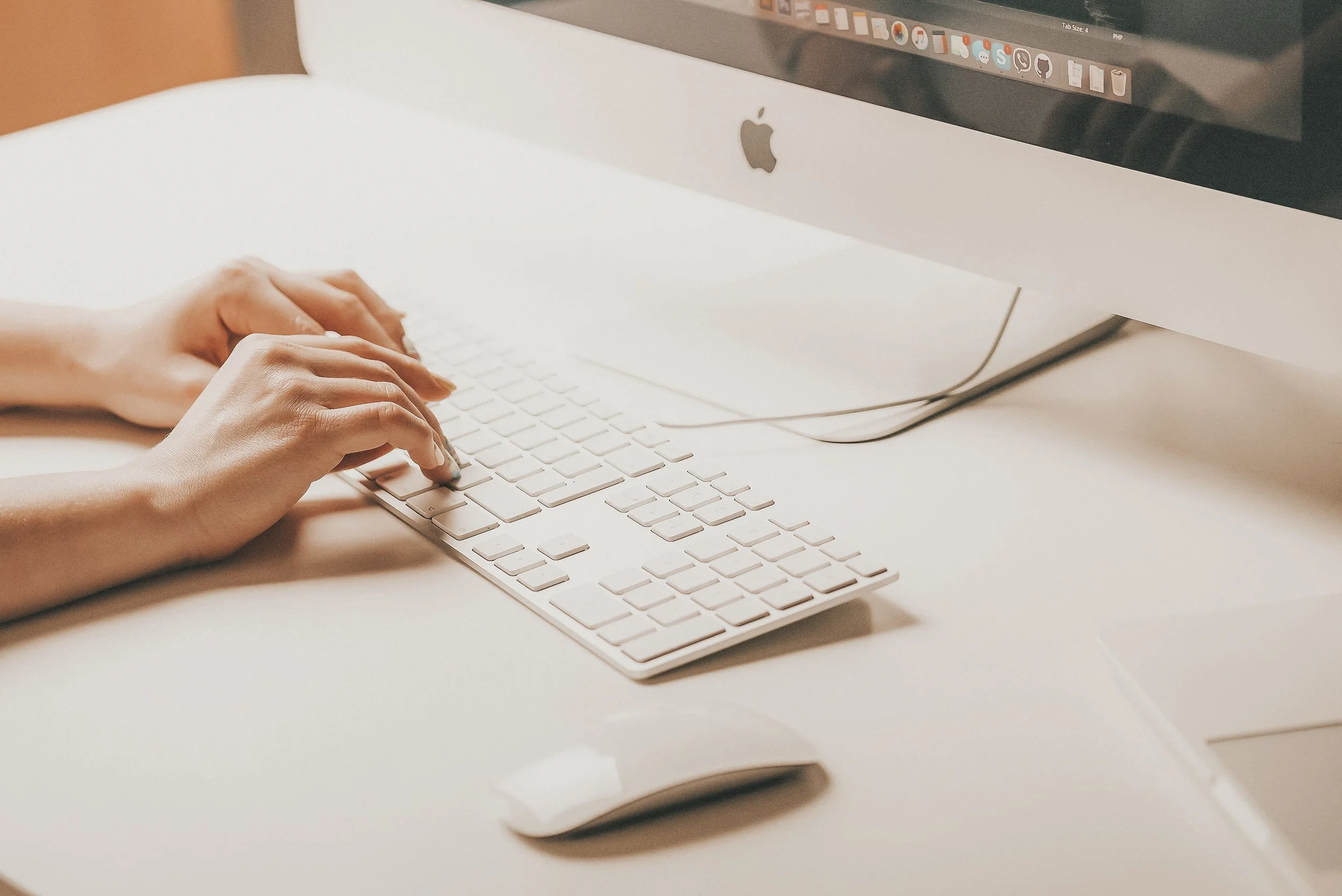 Close-up of person typing on an Apple wireless keyboard and using an iMac computer with a silver mouse nearby on a white desk.
