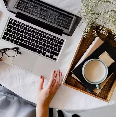 A laptop, a pair of glasses, a hand with red nail polish, a wooden tray holding a mug of coffee, a closed book, a small notepad, and some baby's breath flowers on a white surface.