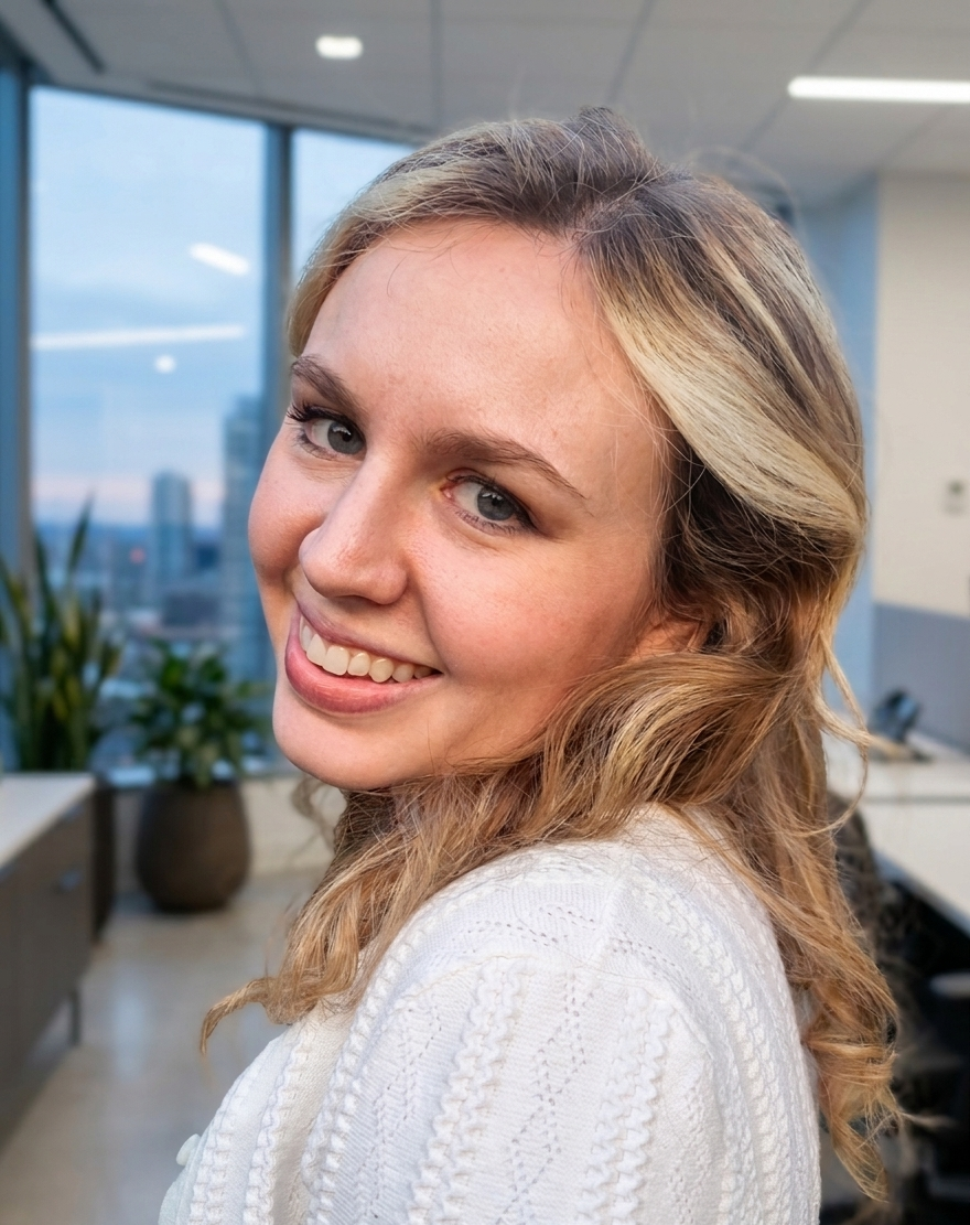 A smiling woman with light brown curly hair and blue eyes in an office setting, with large windows and a cityscape in the background.