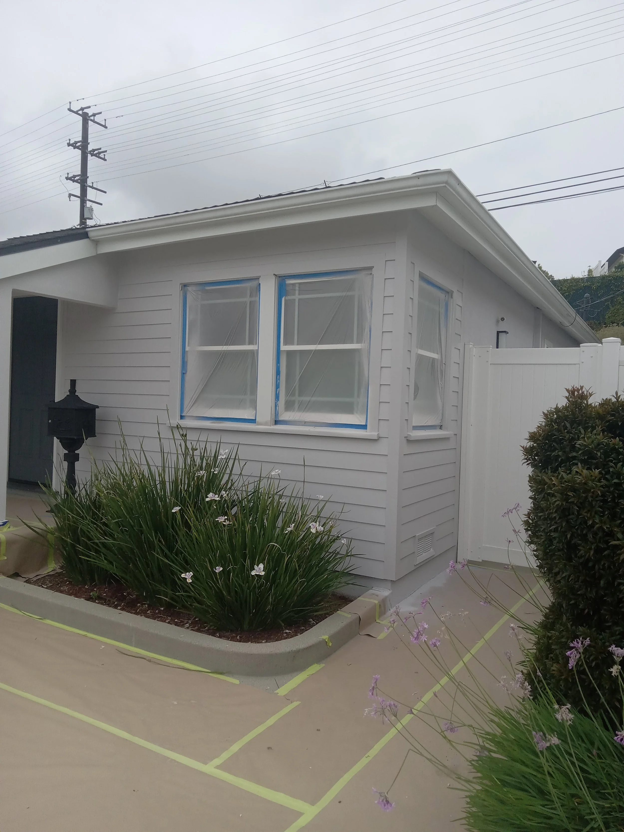 Exterior of a house with white siding, three windows with blue painter's tape, a black mailbox, green bushes, and a concrete sidewalk with yellow markings.
