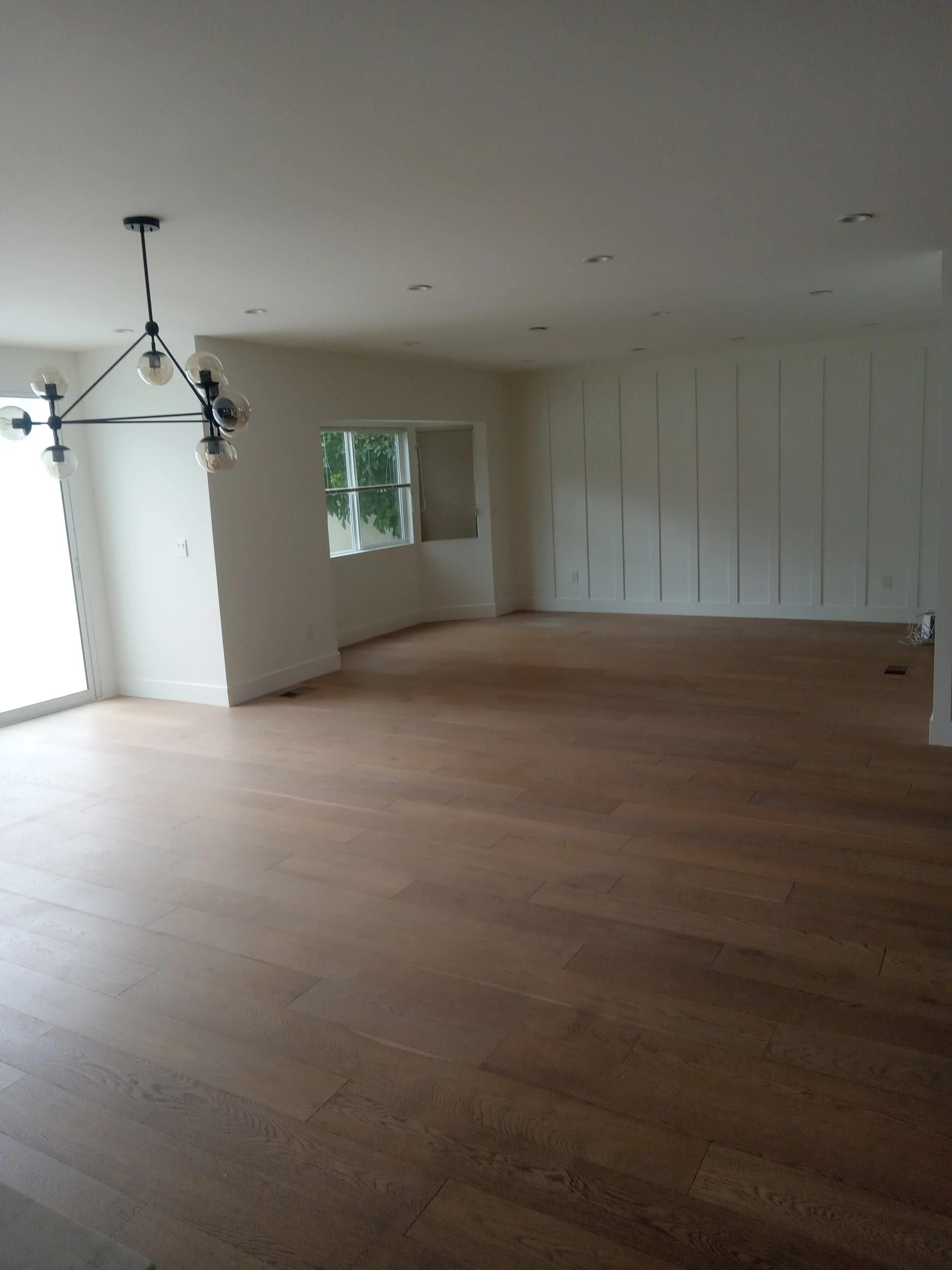 Empty living room with light-colored hardwood floors, white walls, a modern chandelier, and windows allowing natural light.