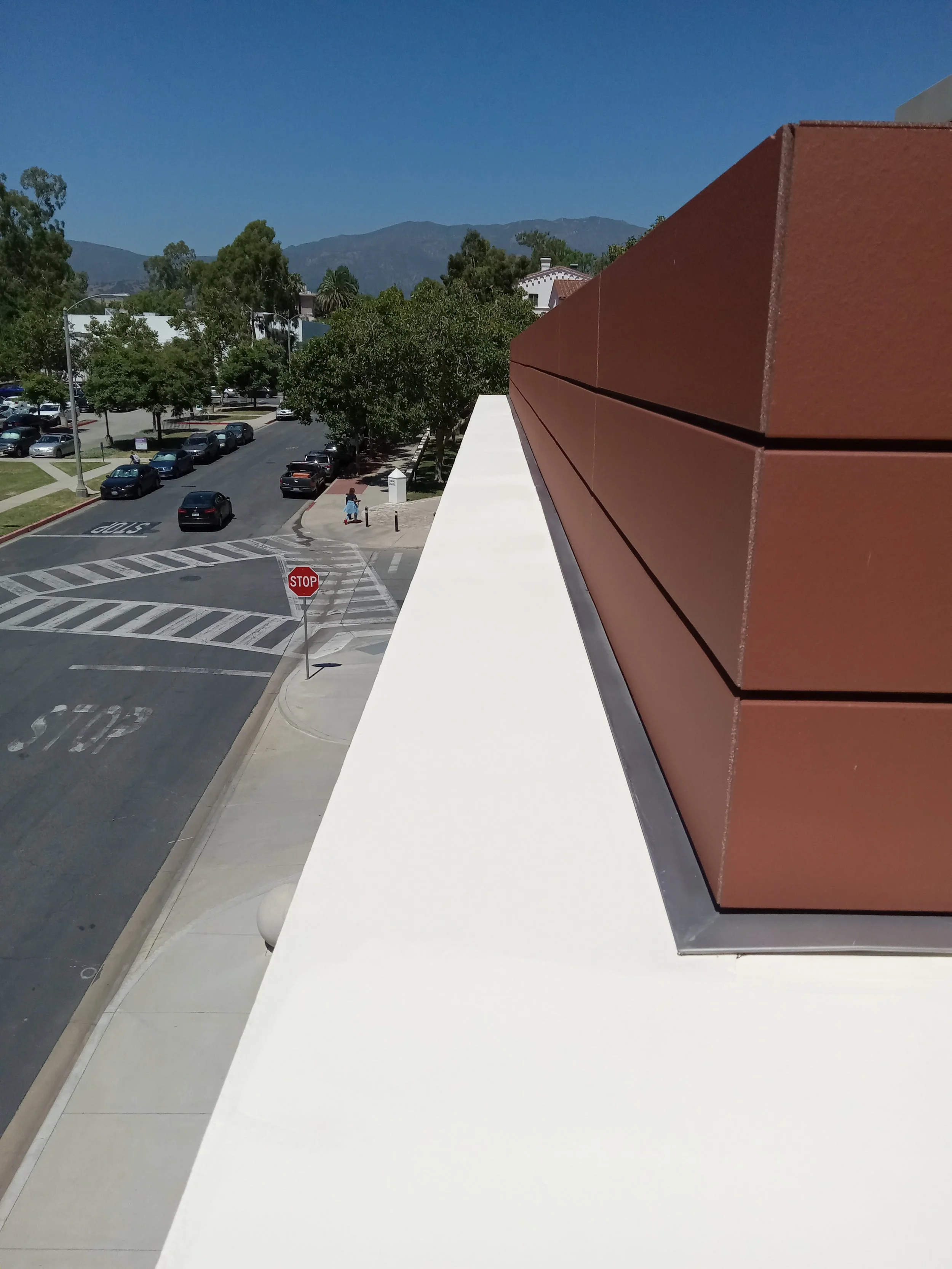 View from a building rooftop showing a crosswalk and stop signs at an intersection, with parked cars, pedestrians, trees, and mountains in the background on a sunny day.