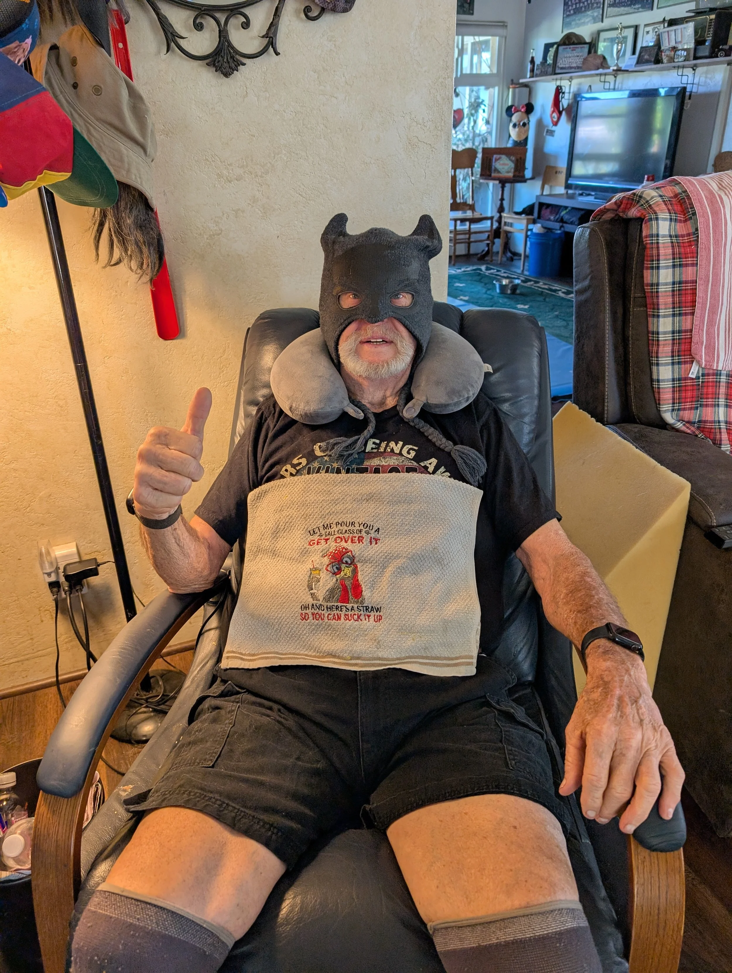 An older man with a gray beard sitting in a recliner in a living room, wearing a black mask with horns, a neck pillow, a graphic t-shirt, black shorts, and knee-high socks giving a thumbs-up.