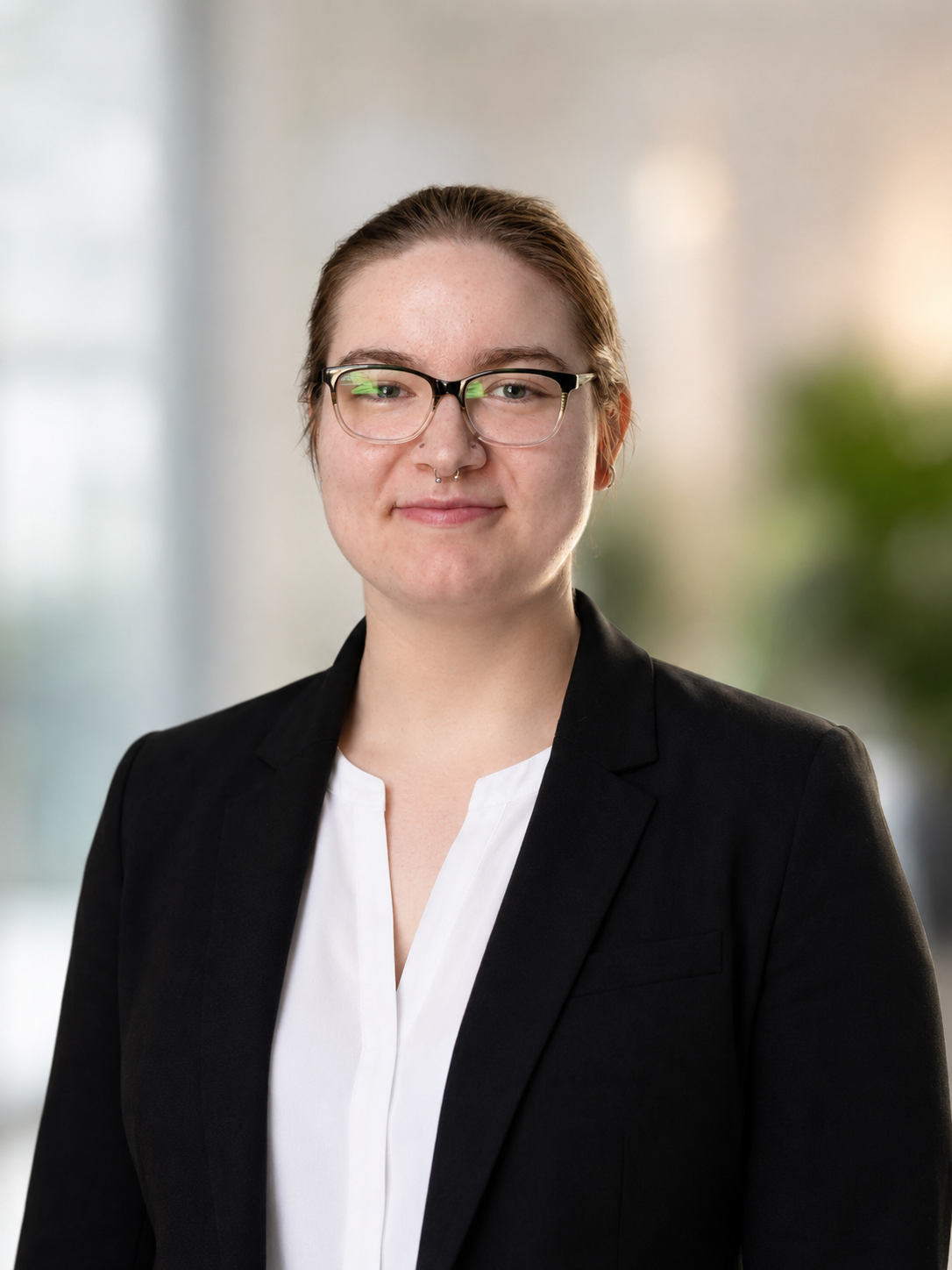 A woman with glasses and a nose piercing wearing a black blazer and white blouse, standing indoors with a blurred background.