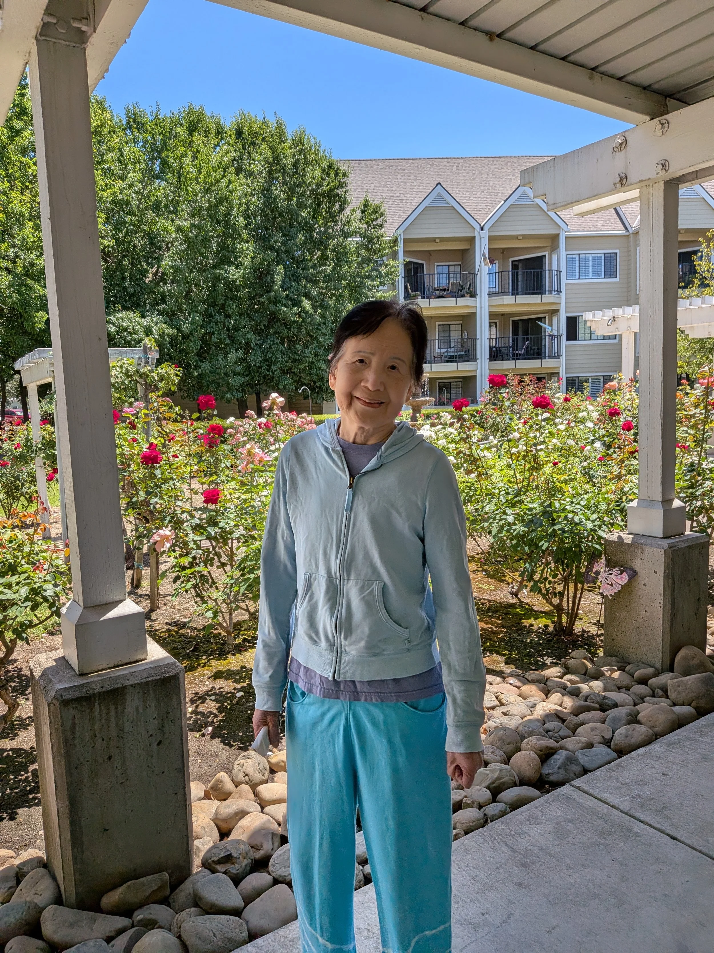 Older woman standing outside on a porch, smiling, with colorful flowers and a beige multi-story apartment building in the background.