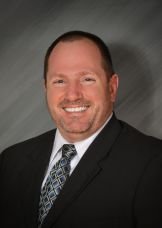 Portrait of a man in a suit smiling against a gray background.