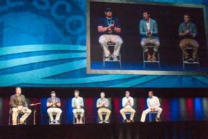 Six men seated on stage in front of a large screen showing the same three men.