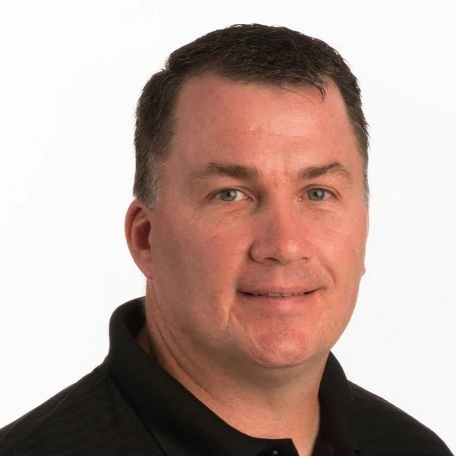 A portrait of a middle-aged man with short brown hair and blue eyes, wearing a black collared shirt, against a plain white background.