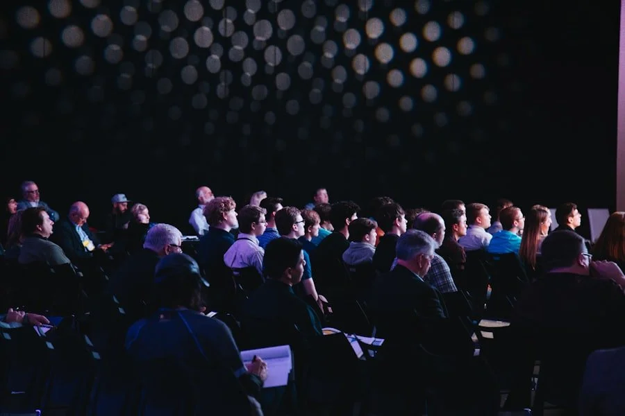 Audience watching a presentation in a dark room with overhead spotlights.