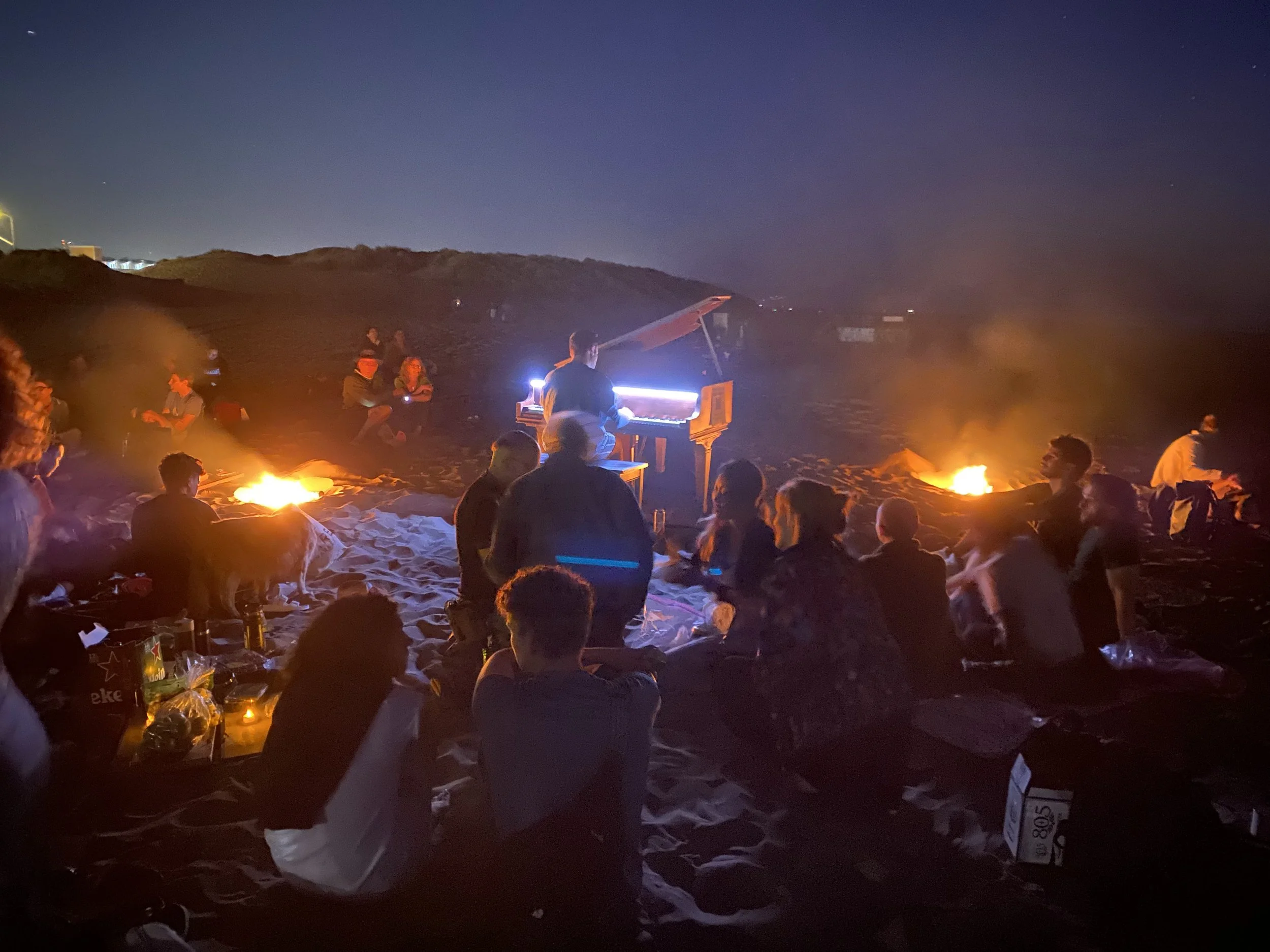 People gathered around a flaming campfire on a beach at night, with a grand piano being played by a musician.