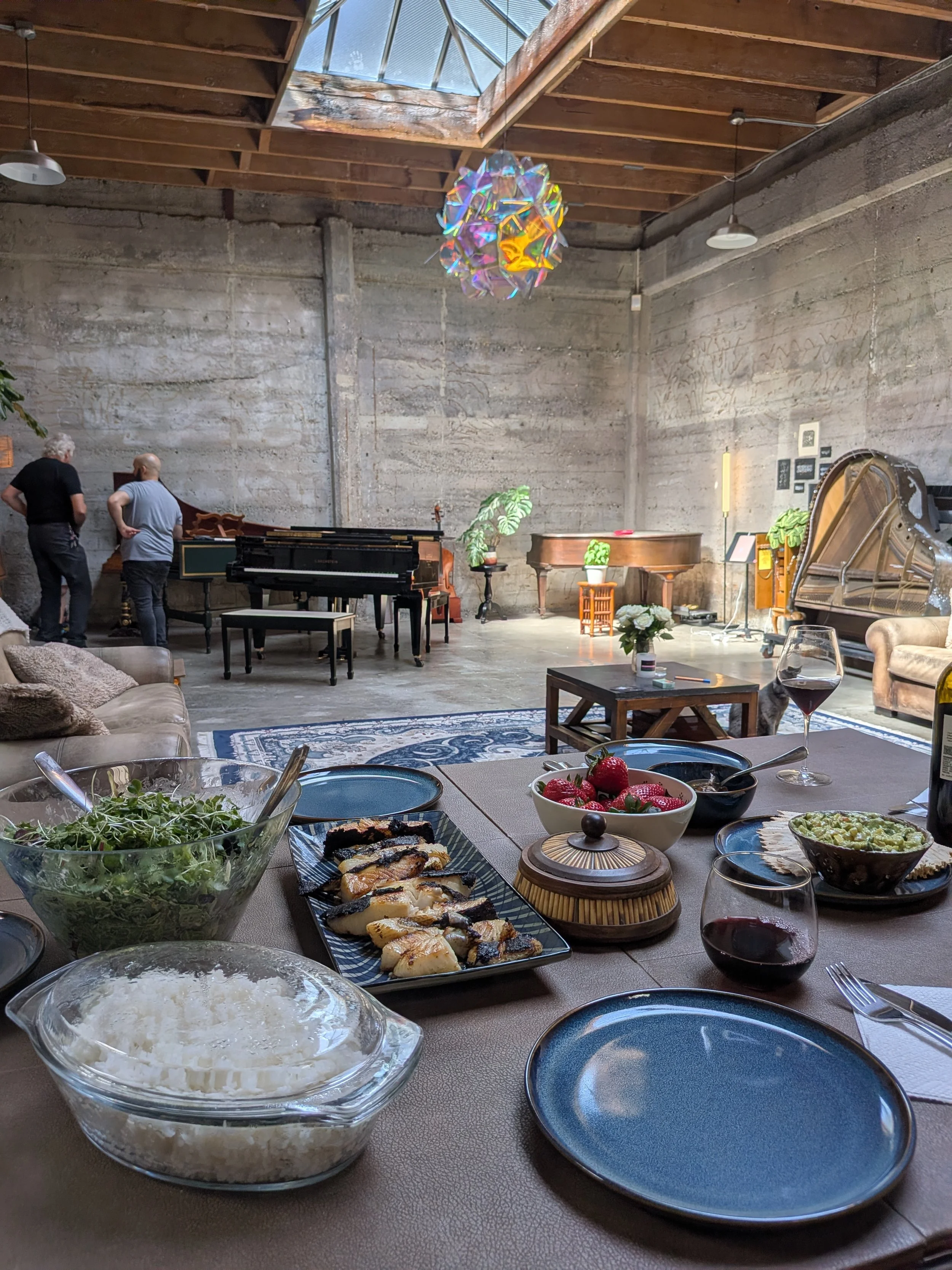 A table set with various dishes including a bowl of strawberries, a bowl of rice, salad, and wine glasses, with a music studio or lounge in the background featuring pianos and a large colorful hanging light fixture.