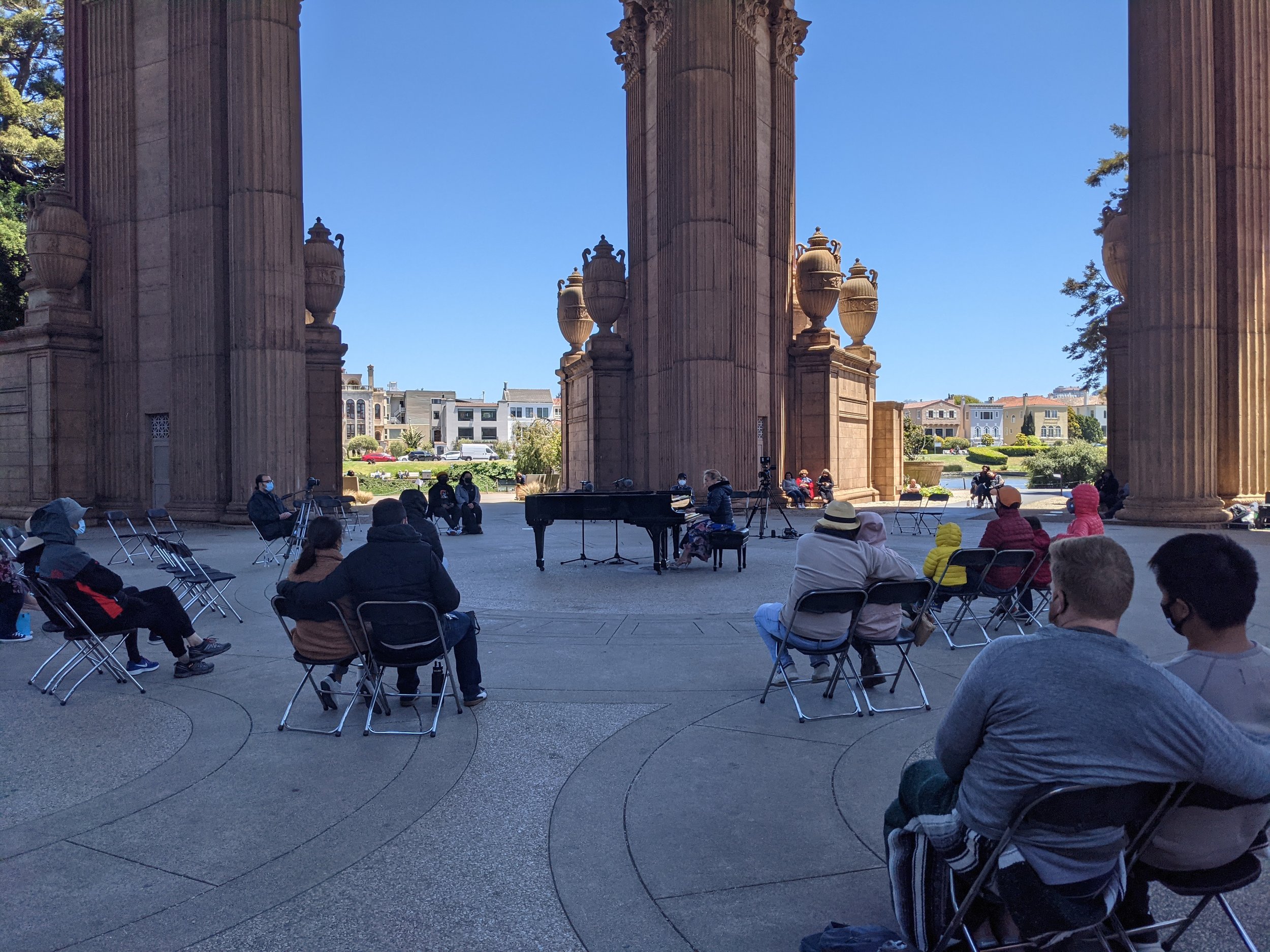 People seated on chairs outdoors, attending a live music performance under large classical columns with a grand piano and a pianist playing, in a park with residential buildings in the background on a clear day.