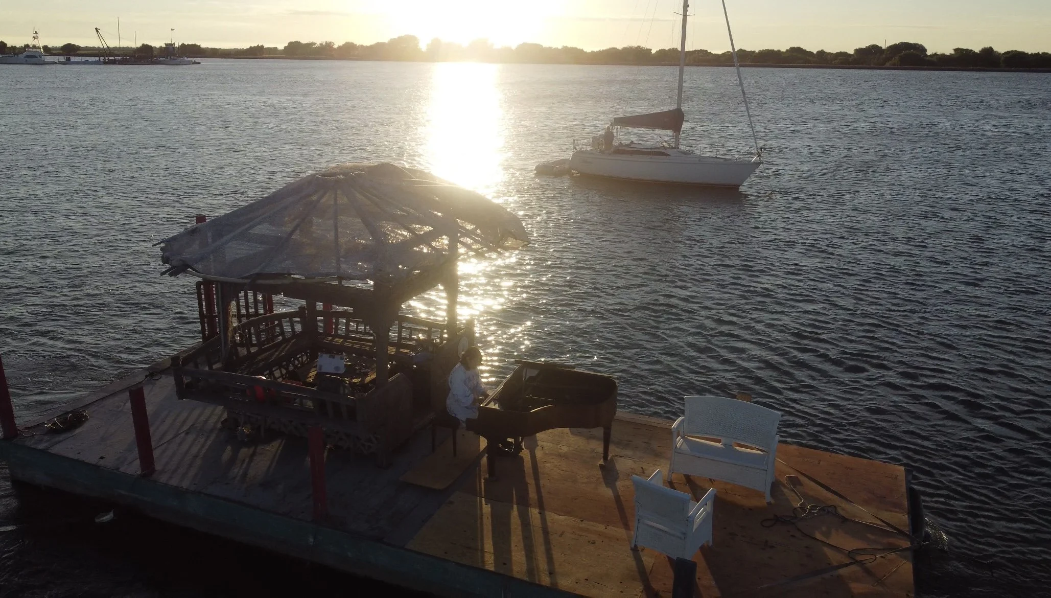 A person playing a grand piano on a floating dock by a body of water with sailboats in the distance during sunset.
