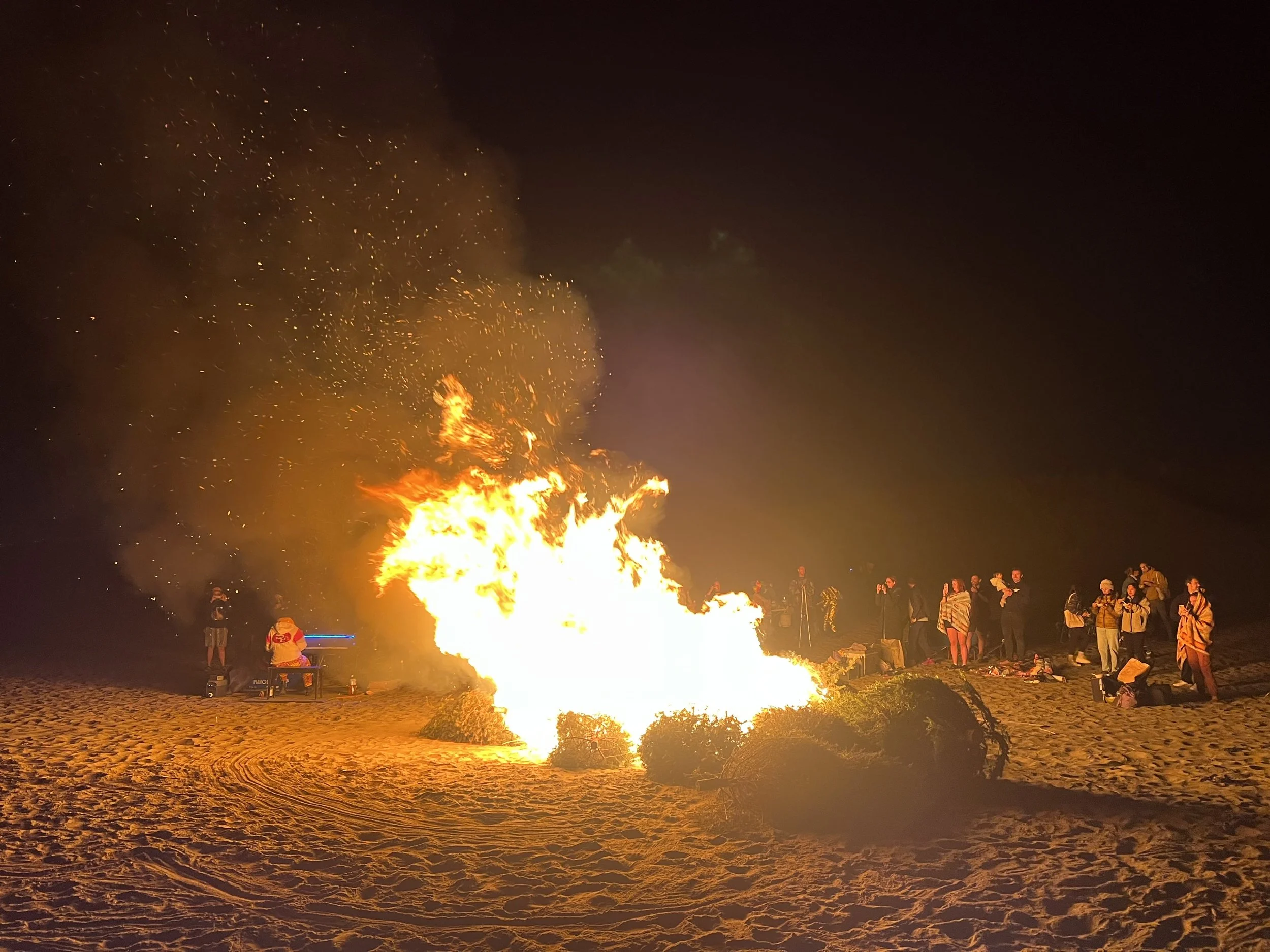 Nighttime scene at a beach with a large bonfire. Several people are gathered nearby, some standing and some sitting, watching the fire.
