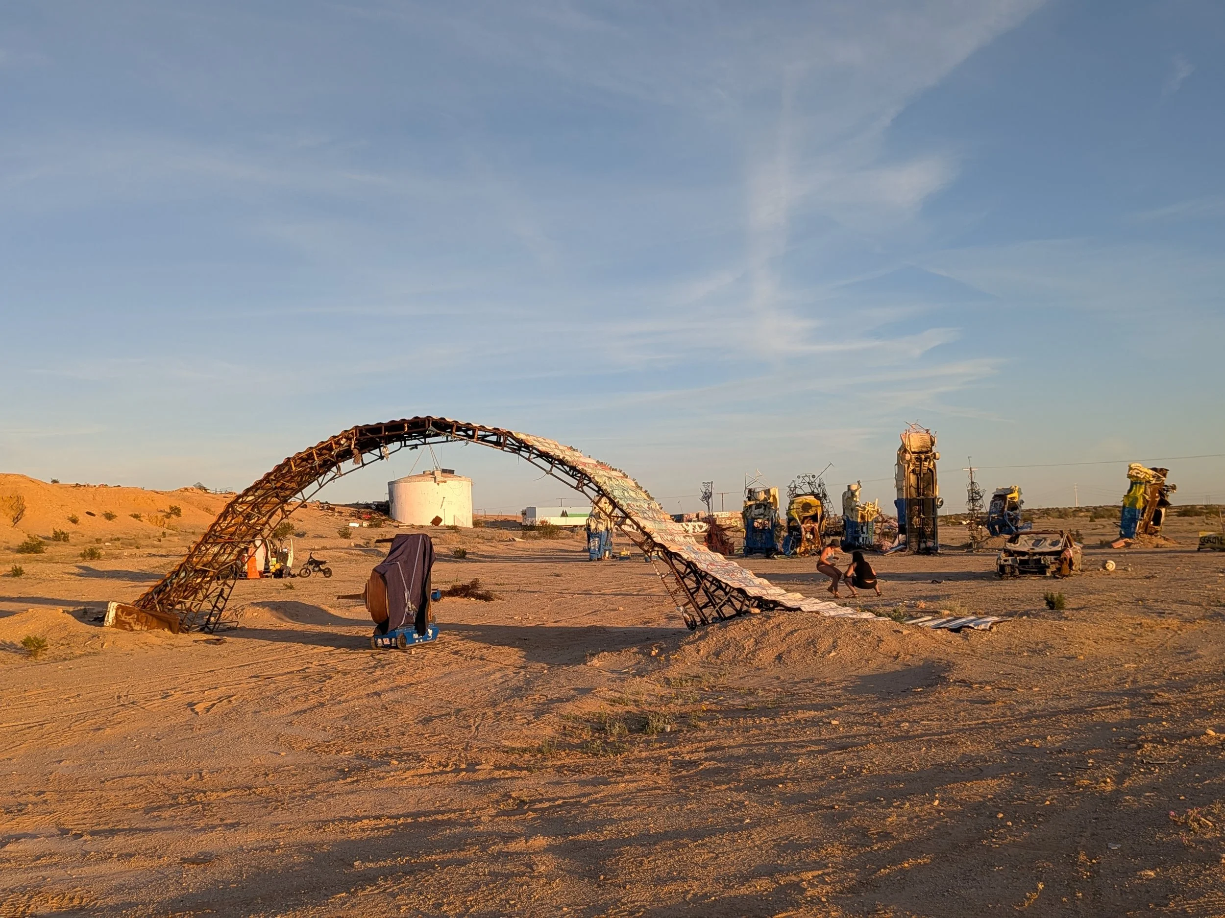 Desert landscape with sculptures resembling mechanical and robotic figures, including a large metal arch and various twisted metal structures, under a clear sky.