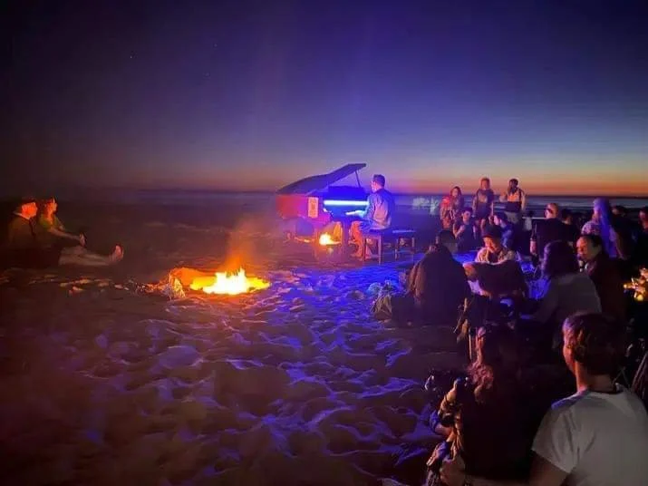 A group of people sitting around a campfire on the beach at sunset, listening to a person playing a piano beside a small fire, with a starry sky overhead.