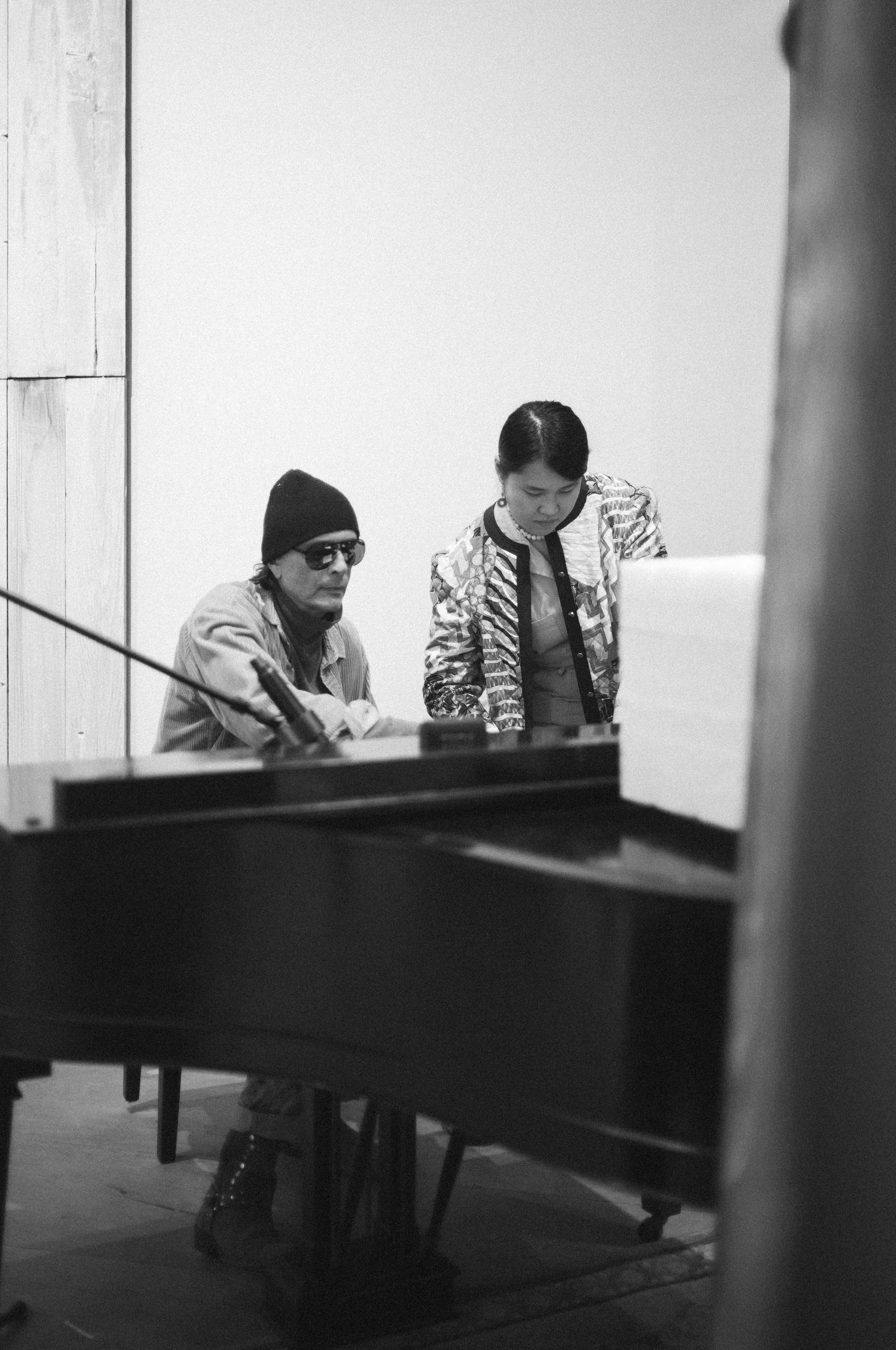 A man wearing sunglasses and a beanie sits at a piano with a woman standing next to him, both looking down at the piano keys in a room with plain walls.