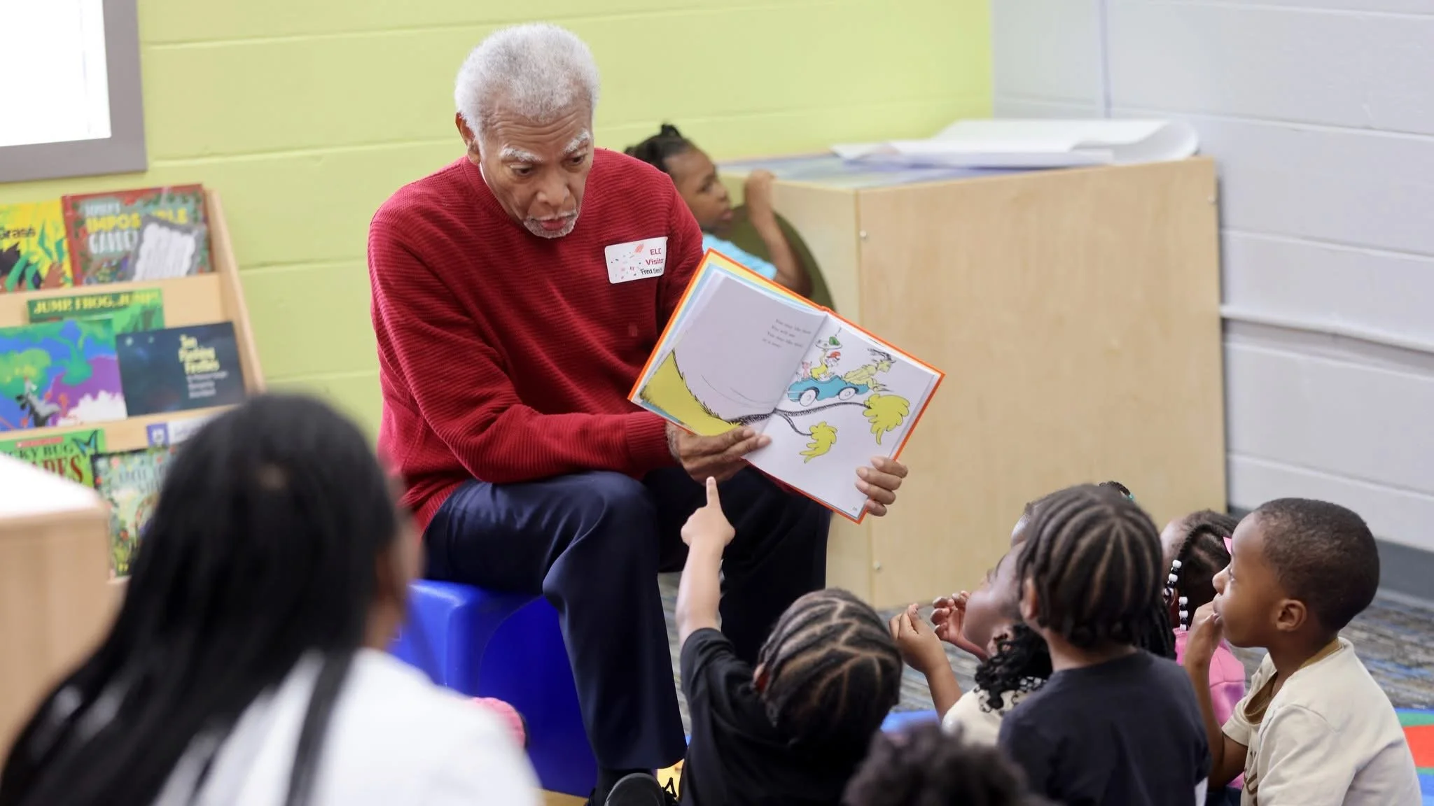 Fred Smith, wearing a red sweater,  reads aloud to children during National Reading Day at the Early Learning Center, sharing a story and engaging young students in a lively classroom reading circle.