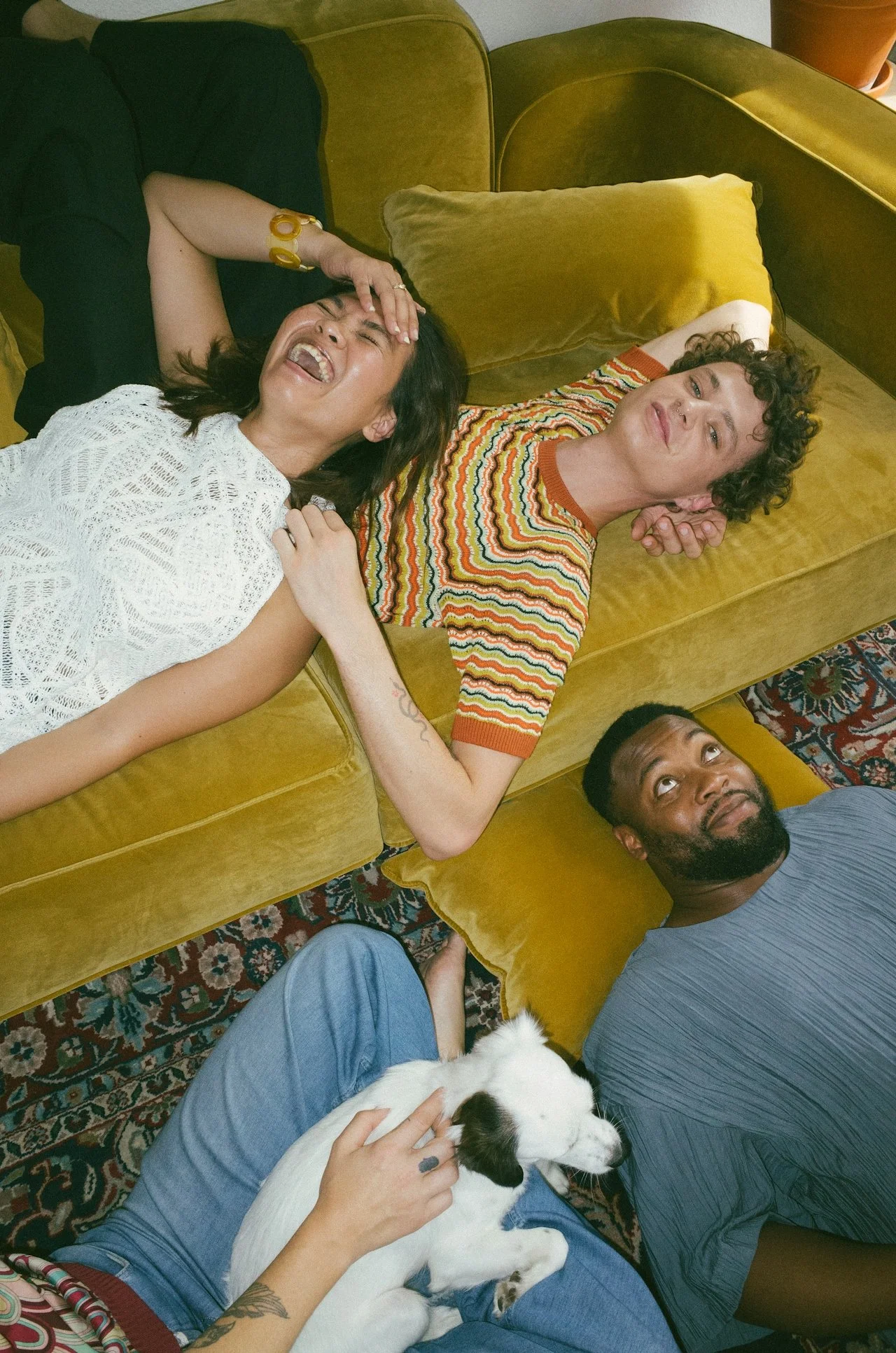 Four people and a dog lying on a mustard yellow couch and a floor with a patterned rug. One person is laughing with hand on forehead, another is looking at camera with a relaxed expression, a third person is gazing upward, and the fourth person is gently petting a white dog with black spots.