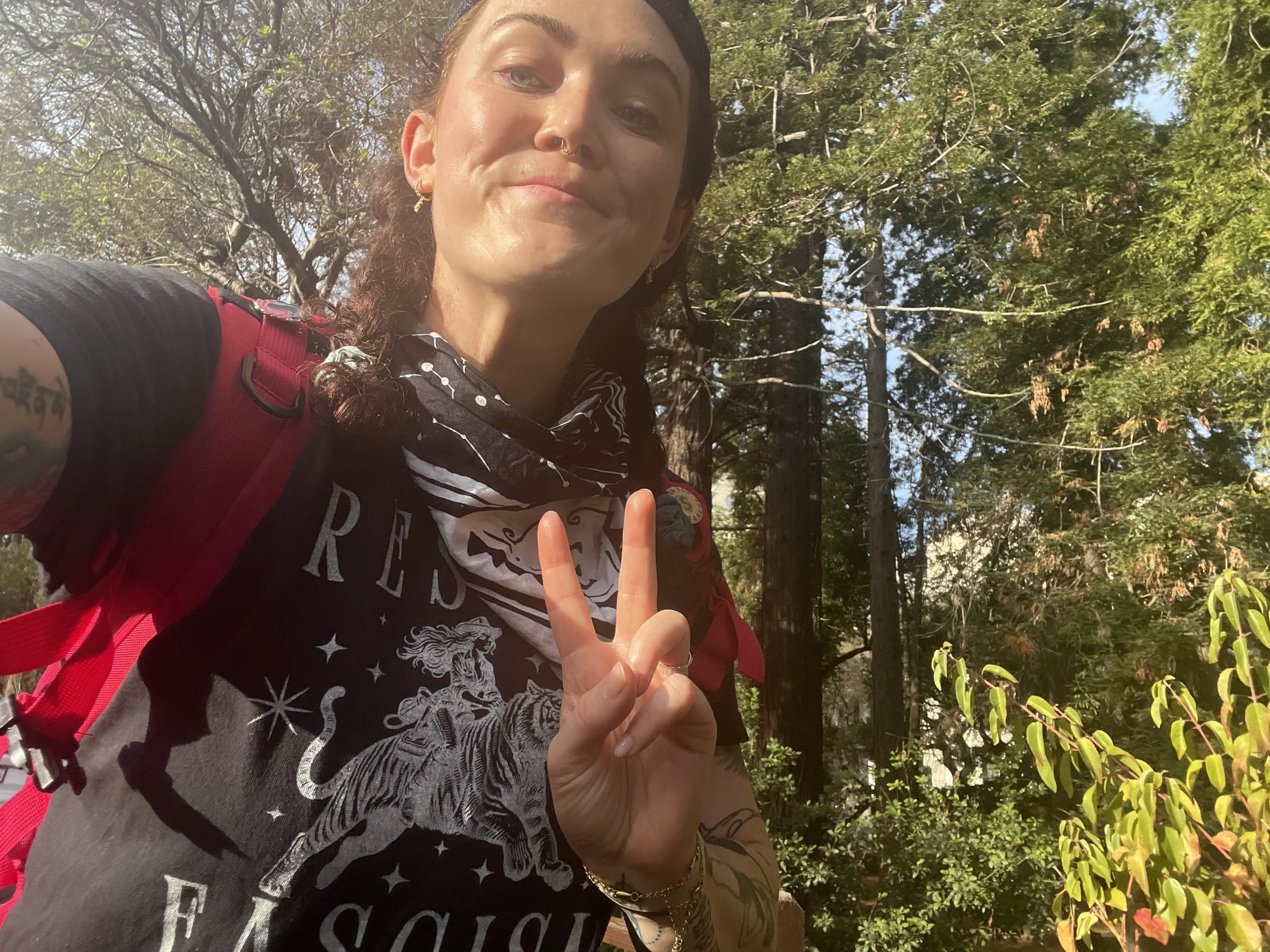 A woman taking a selfie outdoors in a wooded area with green leaves and tree branches in the background. She has tattoos on her arm, is wearing a black shirt with white designs, and a black bandana around her neck. She is also wearing several earrings and has her hair tied back.