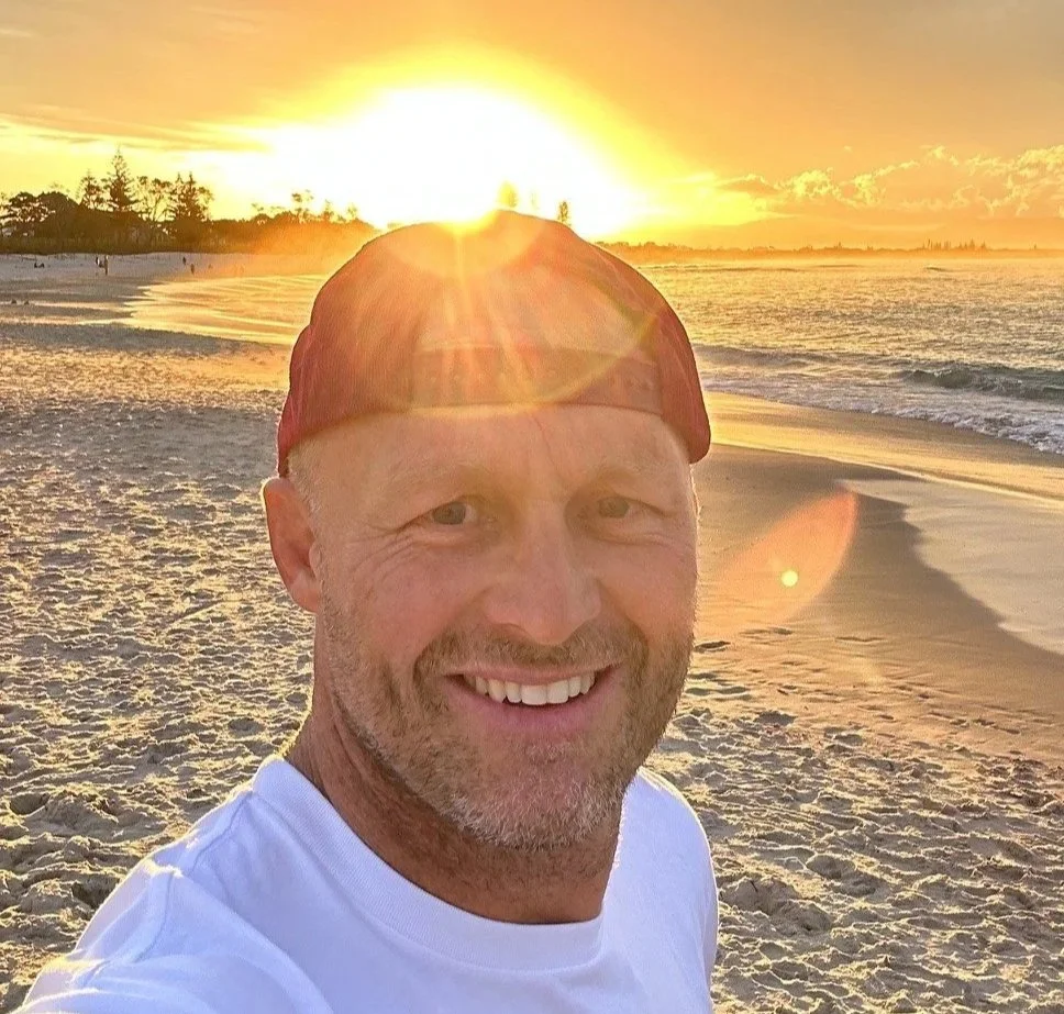 A man smiling on a sandy beach during sunset, wearing a white shirt and a red hat, with the sun setting over the ocean in the background.
