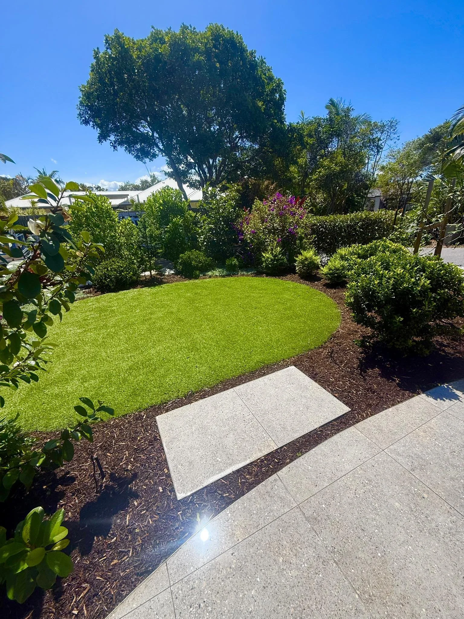 A well-maintained garden with lush green grass, bushes, and a large tree under a clear blue sky. There are concrete pavers bordering the garden and a paved area nearby.