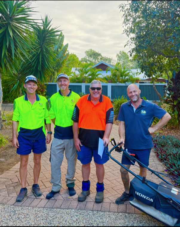 Four men standing outdoors on a brick pathway in front of lush greenery and a blue fence, smiling and dressed in colorful work uniforms, one of whom is holding a lawn mower.