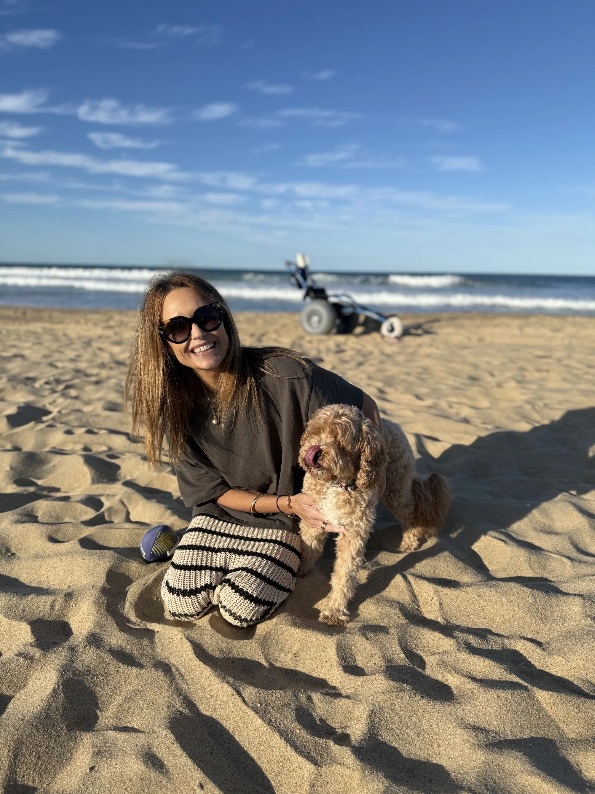 A woman with long hair and sunglasses smiling while sitting on the sand on a beach, holding a curly-haired dog. The ocean waves and a beach scooter are visible in the background under a blue sky with some clouds.