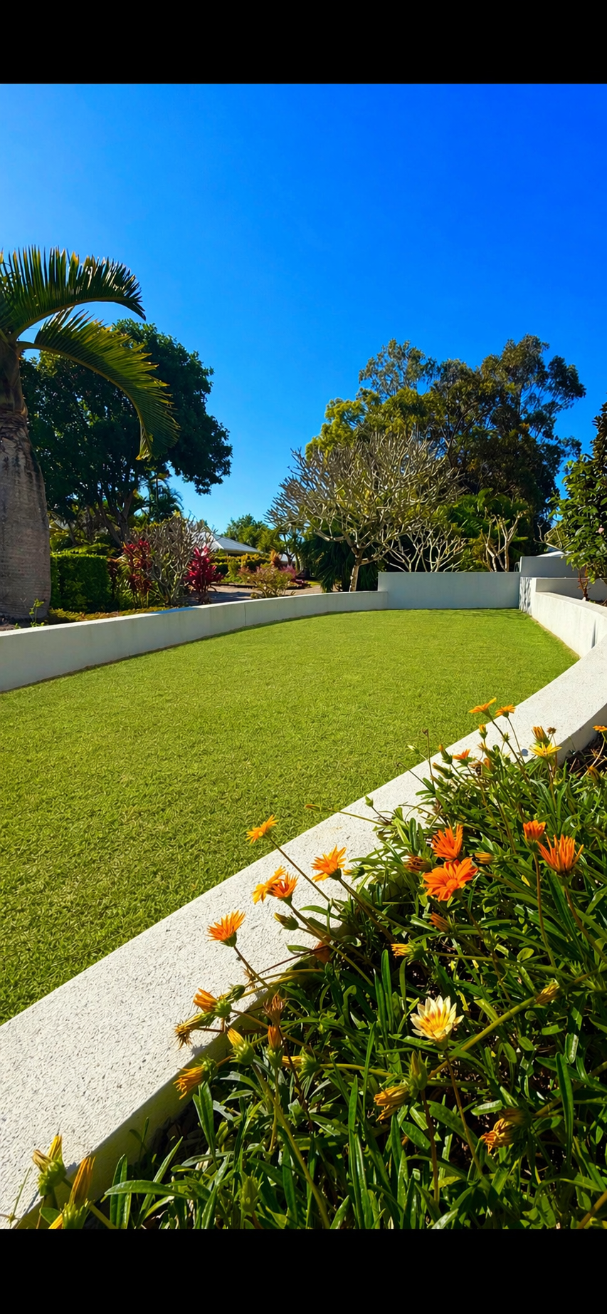 A lush green lawn bordered by white concrete walls, with colorful flowers in the foreground, tropical trees, and a clear blue sky in the background.