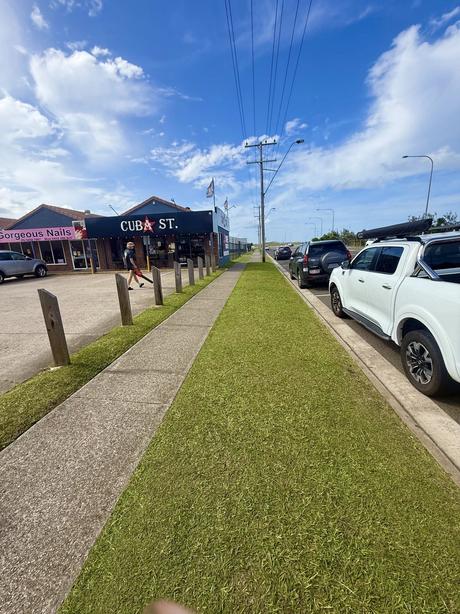 Street view showing a sidewalk, parked cars, utility poles, and a building with signs for a nail salon and a bar called 'Cuba St'. The sky is partly cloudy with a bright, sunny day.