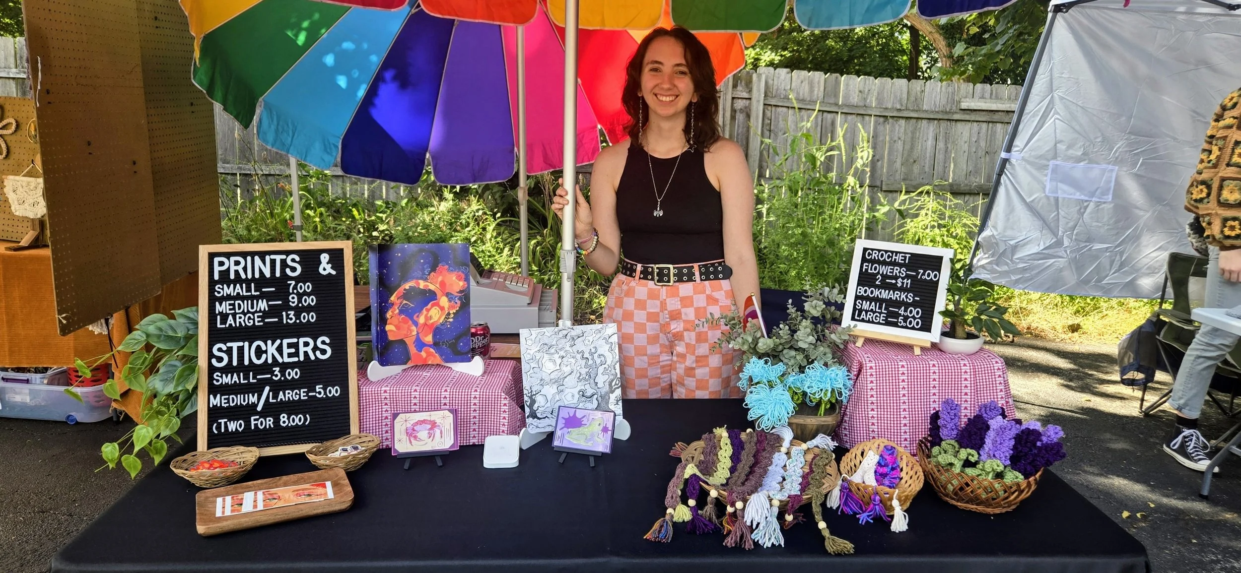 A young woman standing behind a market stall selling crocheted items, art prints, stickers, and craft flowers, with a multicolored umbrella overhead at an outdoor market.