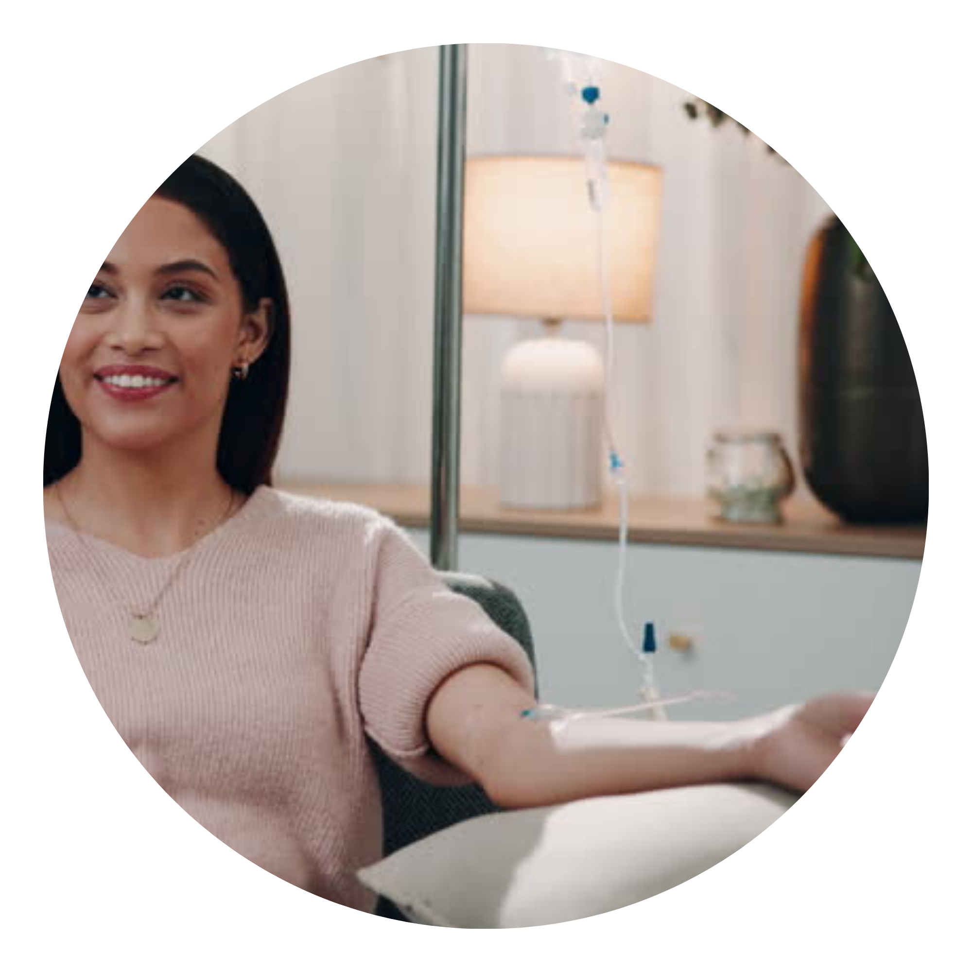 A woman smiling with dark hair wearing a light pink sweater, seated near an IV drip in a clinical setting with a white lamp and medical equipment in the background.