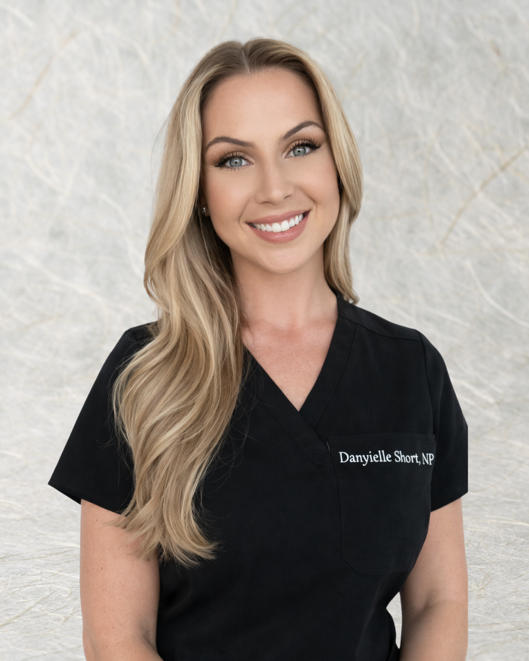 A woman with long blonde hair, blue eyes, and a bright smile wearing a black medical scrub top with the name tag 'Danyielle Short, NP'.