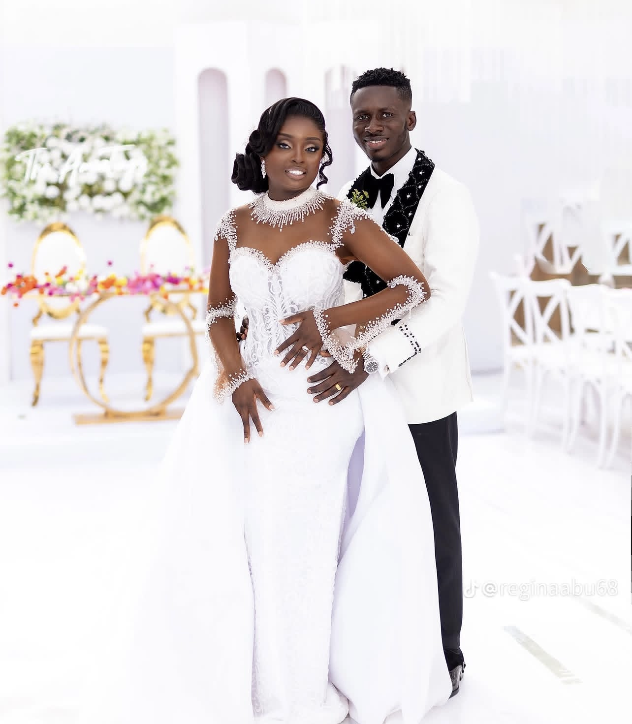 A bride and groom standing together at their wedding, both dressed in formal wedding attire, in a decorated indoor venue.