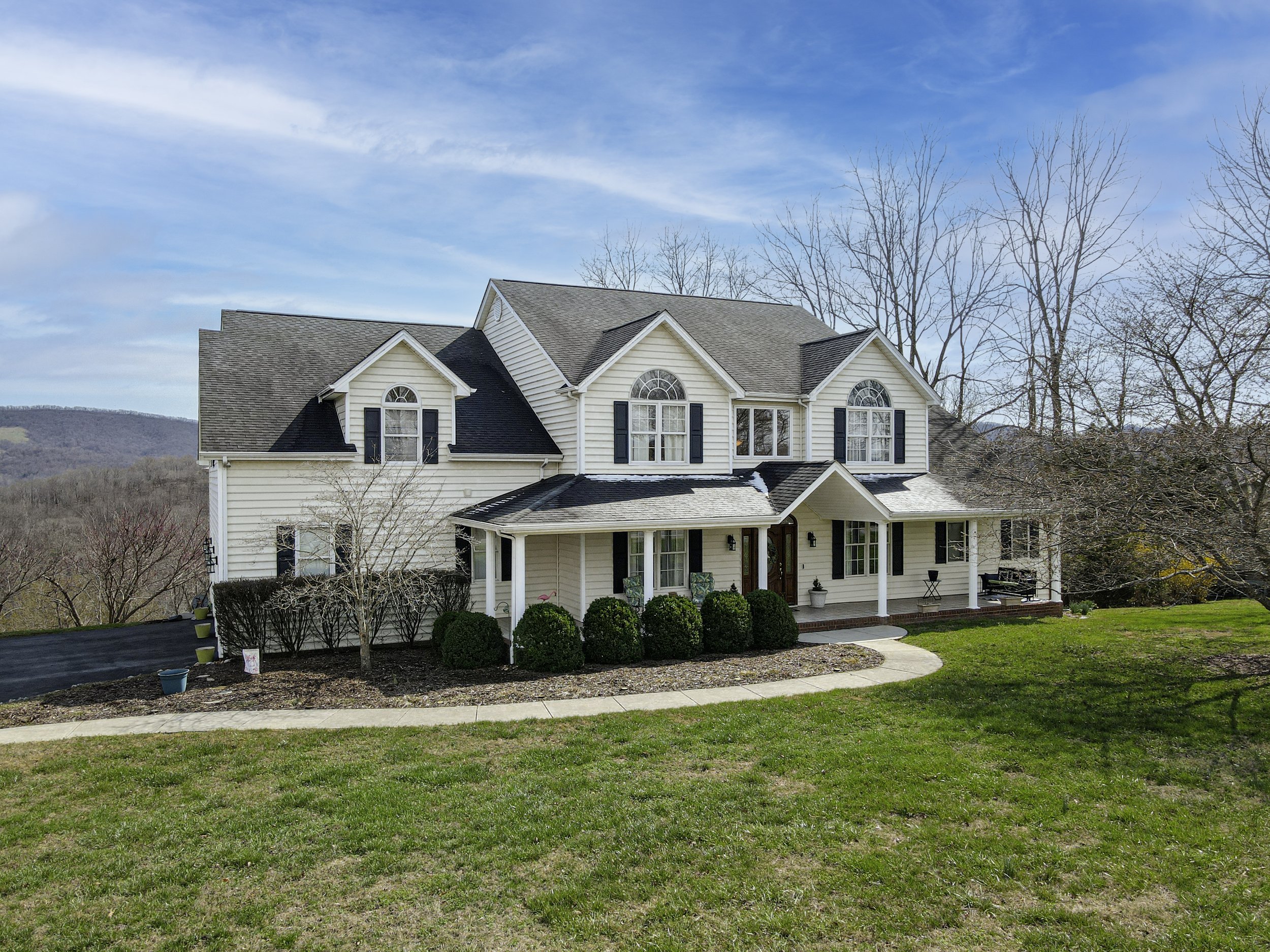 A large, white two-story house with black shutters, a covered front porch, and a landscaped yard amidst leafless trees with a mountainous background under a blue sky.