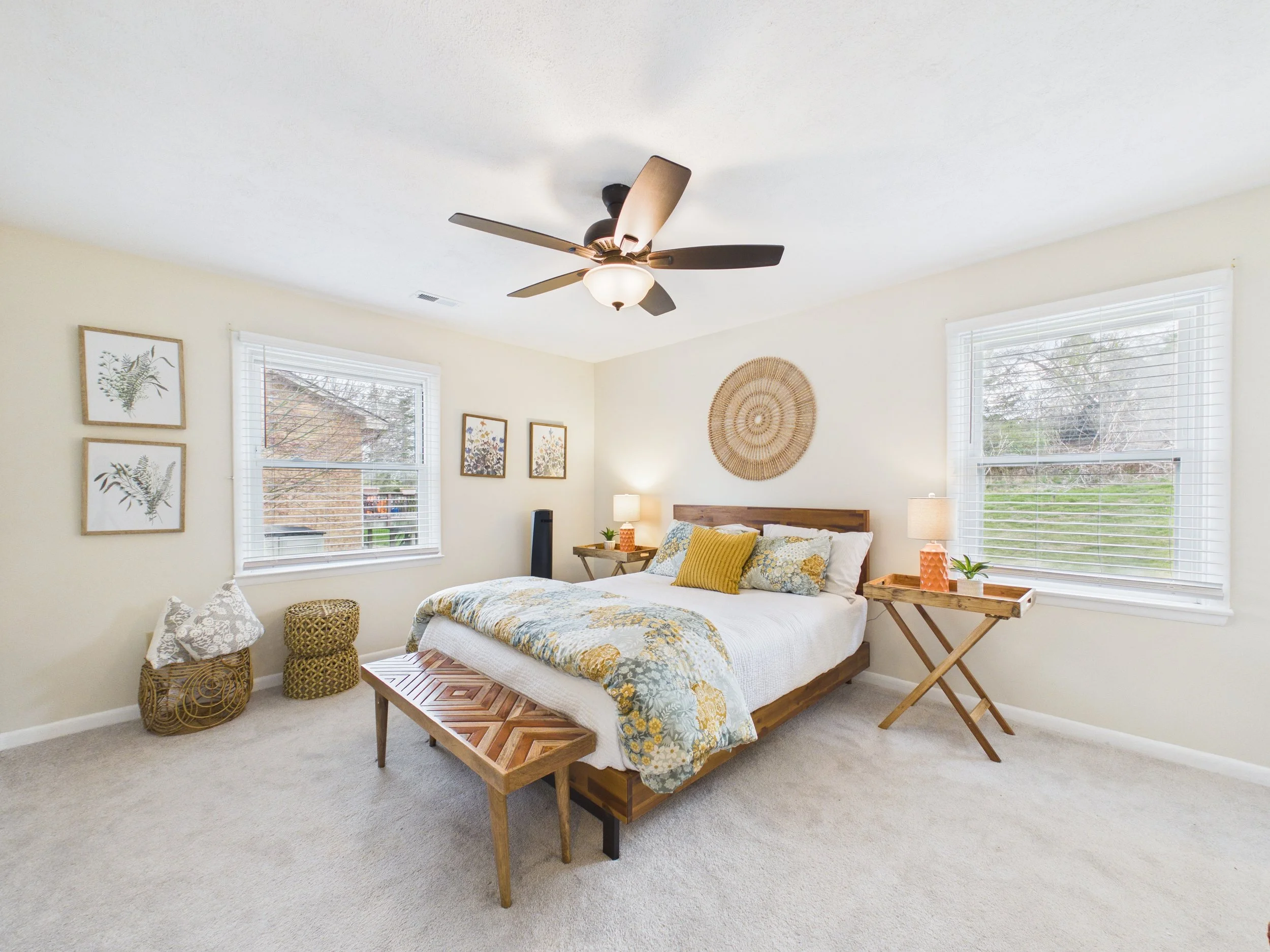 Bright bedroom with a wooden bed, floral bedding, yellow throw pillow, two wooden nightstands with lamps, large windows with white blinds, framed botanical art, wicker basket, decorative wall hanging, ceiling fan, and light-colored carpet.
