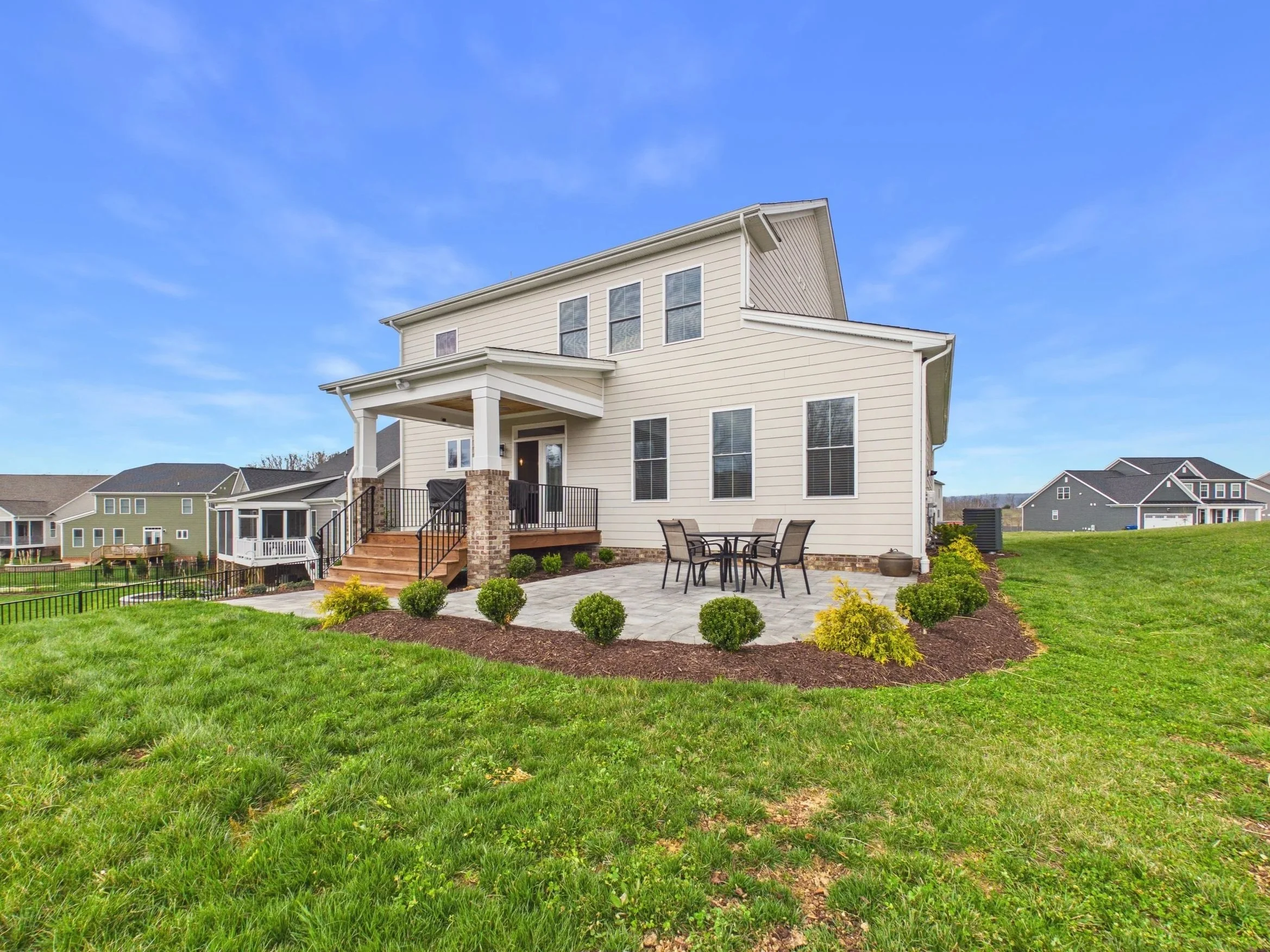 A two-story cream-colored house with a porch and outdoor seating area, surrounded by green grass and landscaping, under a blue sky.