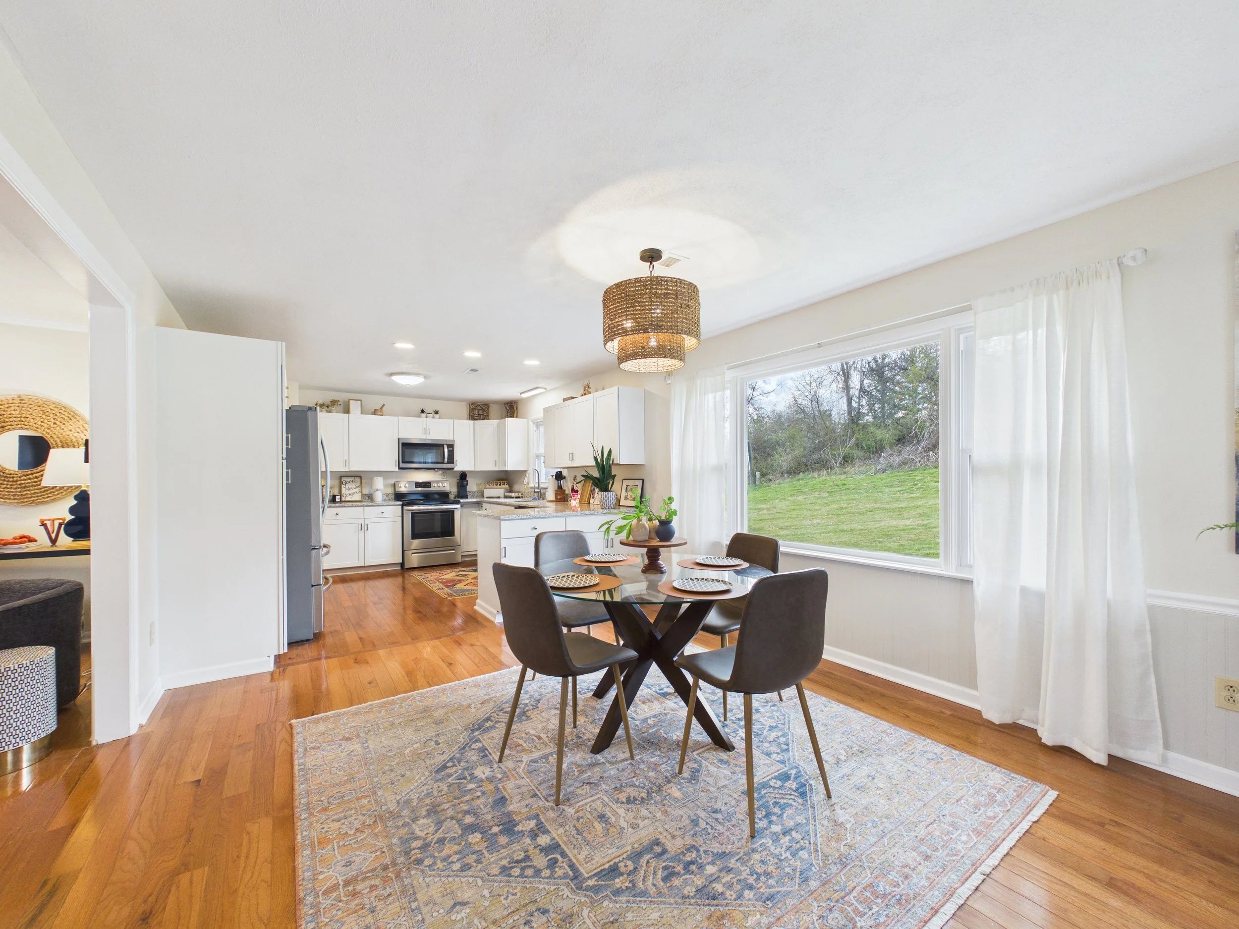 Open-concept dining area and kitchen with hardwood floors, a round dining table with four chairs, a large window with white curtains showing a grassy yard, white cabinets, stainless steel appliances, and a woven pendant light.