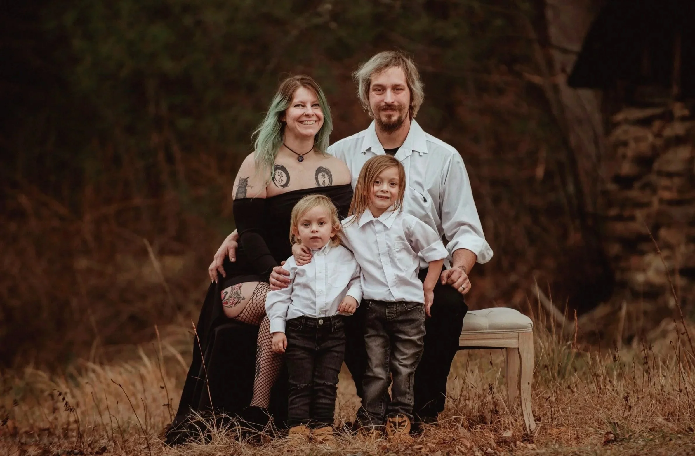 A family of four posing outdoors at sunset, with a background of trees and rocks. The mother has green hair, tattoos, and is wearing a black off-the-shoulder dress. The father has a scruffy beard and is dressed in a white shirt. Their two young children are standing in front, both wearing white shirts and dark pants.