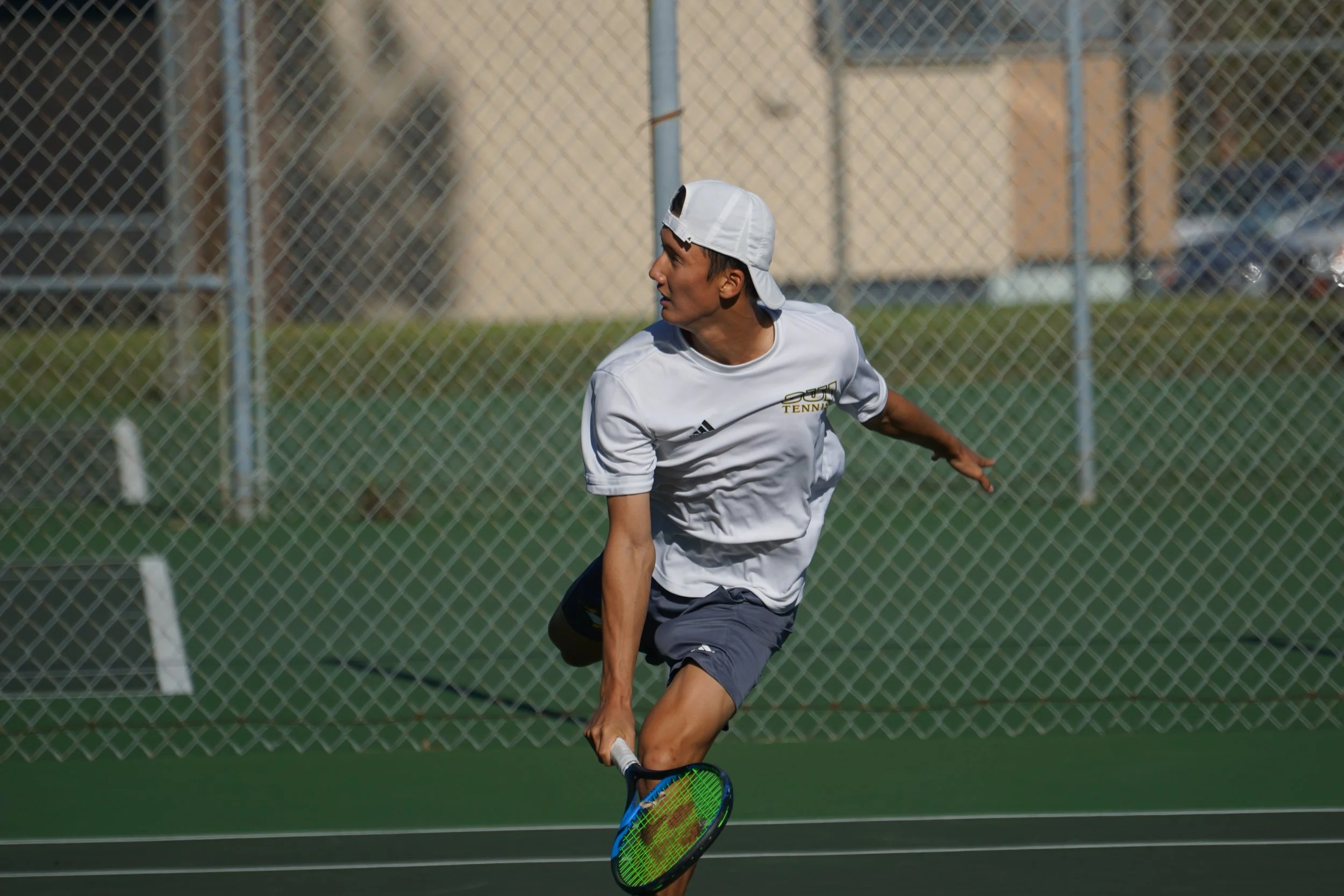 Young man playing tennis on an outdoor court, wearing a white cap backwards, white shirt with 'TENNIS' logo, and black shorts, holding a tennis racket.