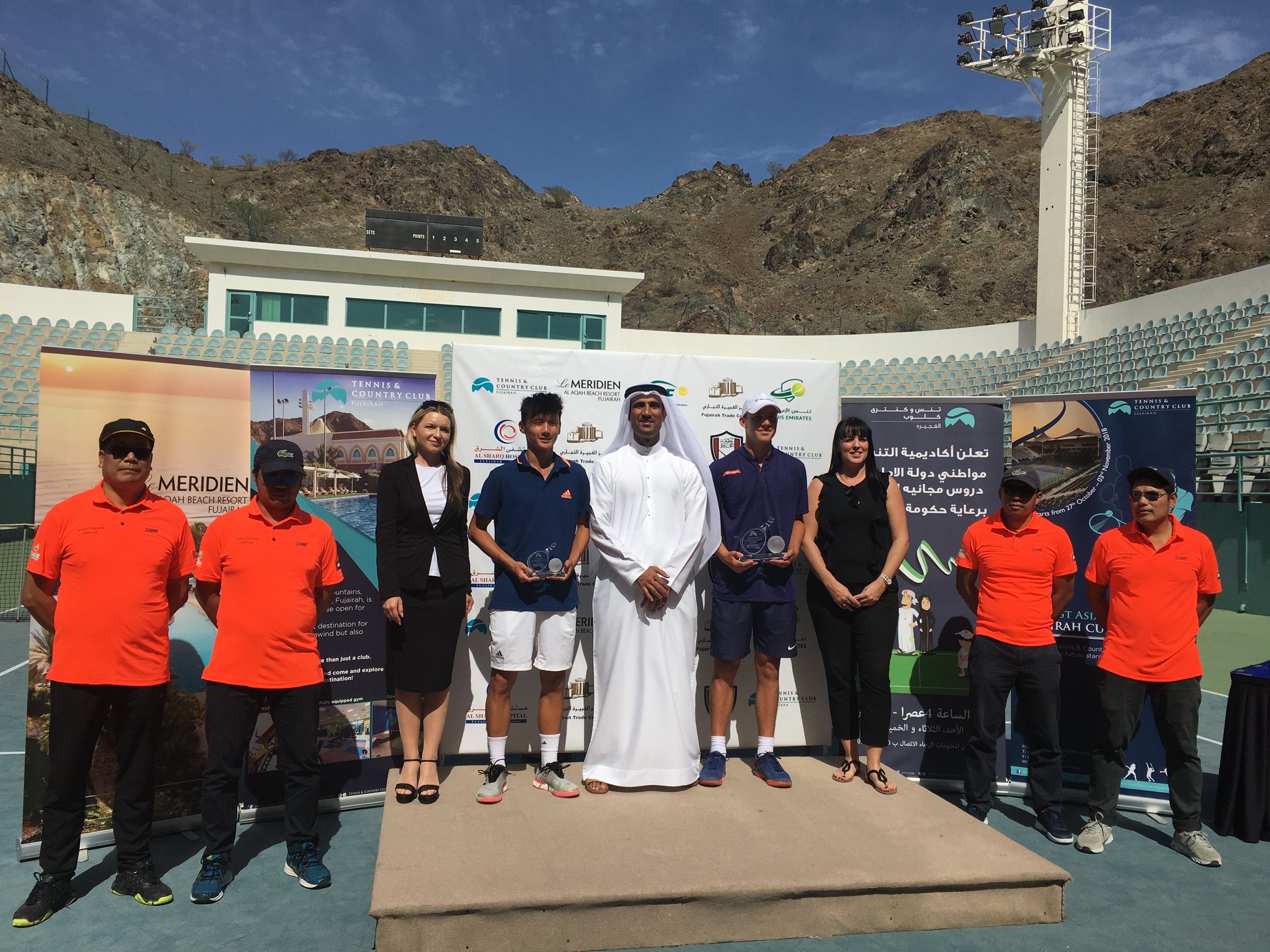 Group of nine people standing on a podium at a tennis event, with a mountainous background and clear sky. Four of them are holding trophies, and banners and signs related to the event are visible behind them.
