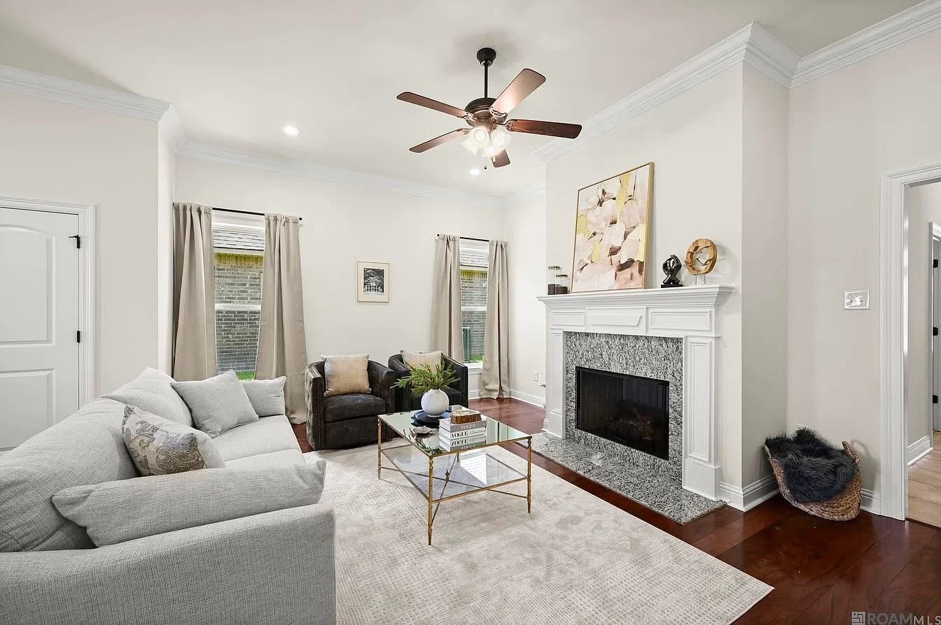 Bright living room with white walls, beige curtains, a white sofa, and two dark armchairs arranged around a glass coffee table. A fireplace with a gray granite surround is decorated with artwork and small sculptures. Hardwood floors and a ceiling fan with light fixtures are also visible.