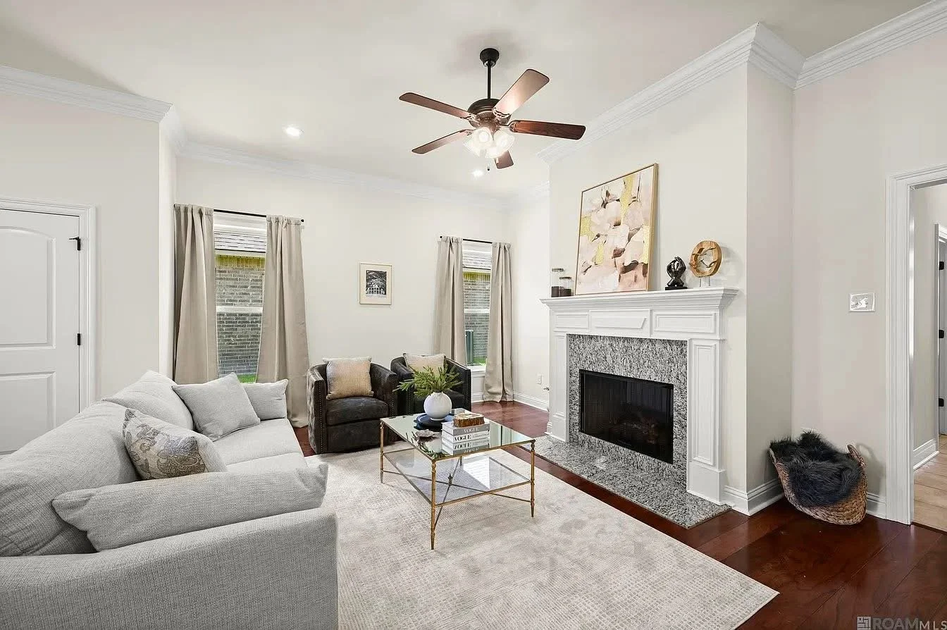 Living room with white sofa, black armchairs, glass coffee table, fireplace with white mantel, hardwood floors, ceiling fan, and beige curtains.