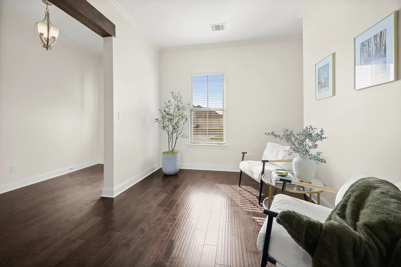 Bright living room with white walls, dark wood flooring, a window with blinds, a beige couch with black armrests, a white armchair, a small side table with books and a plant, and framed pictures on the wall.