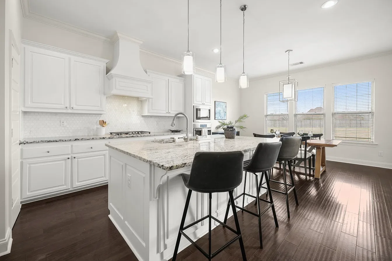 Bright, modern kitchen with white cabinetry, granite island, dark wood floors, and large windows with blinds, featuring bar stools and a dining table.
